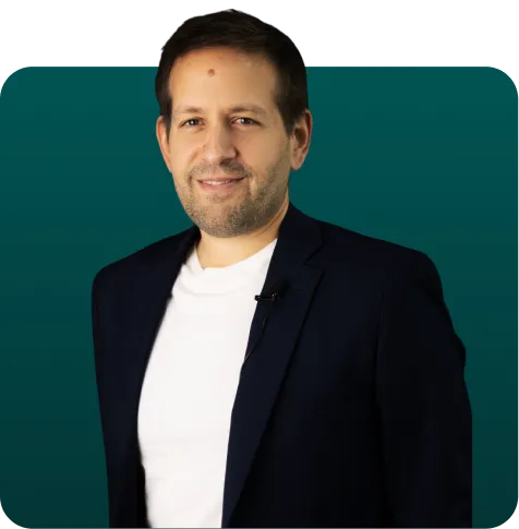 Man with short brown hair and slight beard wearing a white T-shirt and dark blazer against a teal background.