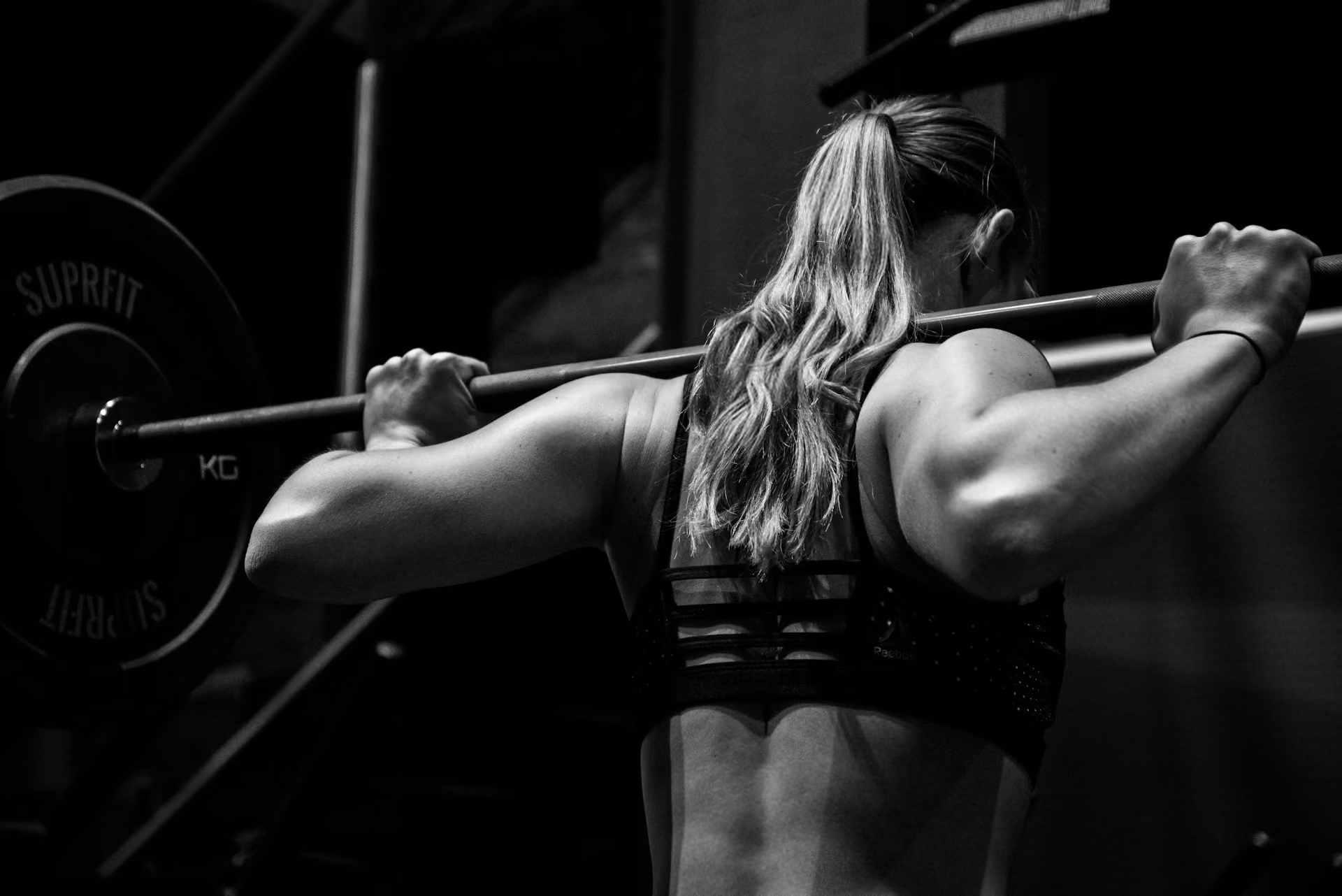 A woman in a gym stands with a barbell on her shoulders, mid lift.