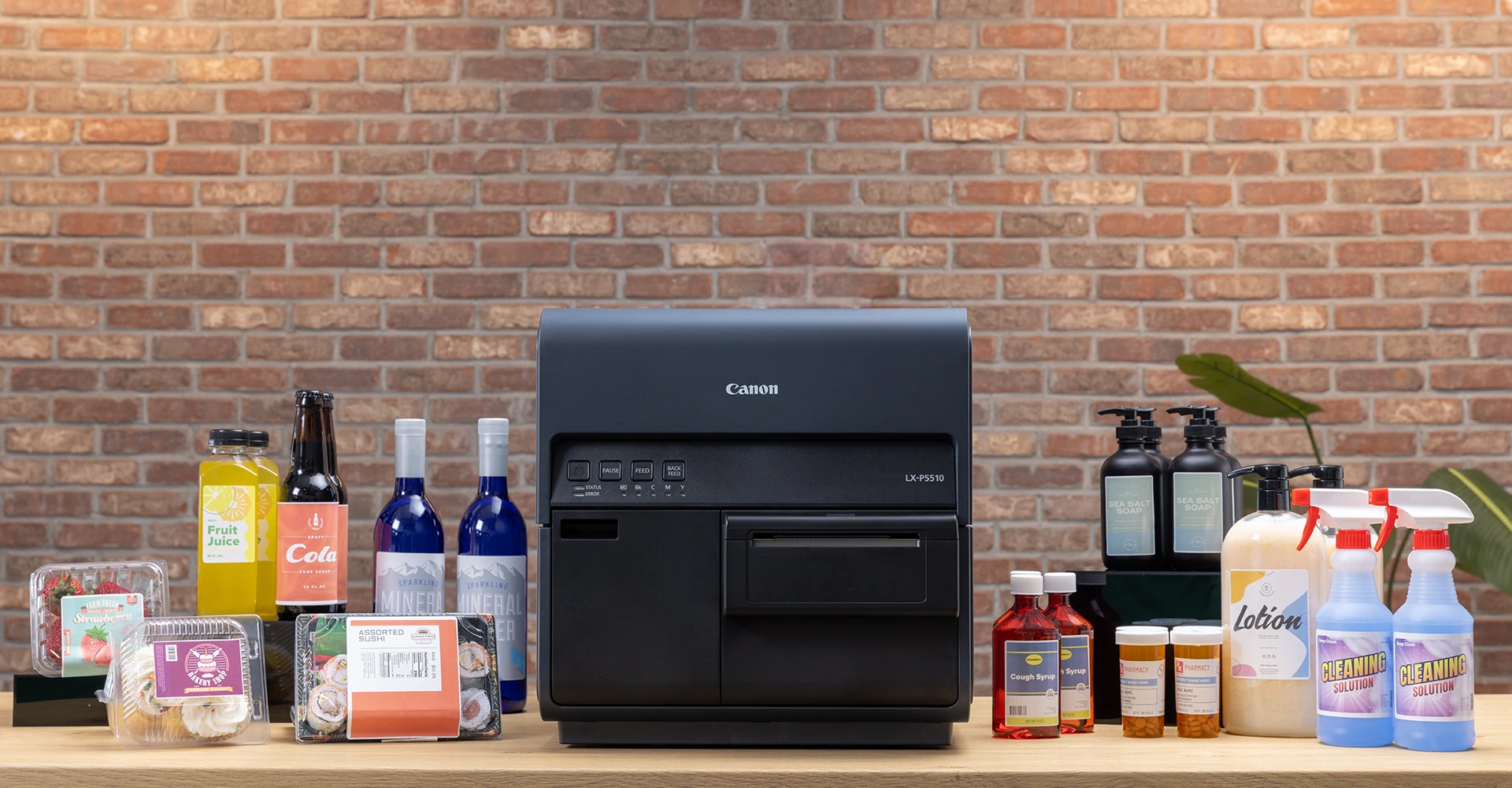 A Canon LX-P5510 colour label printer on a shop counter, surrounded by own brand packaged goods with freshly printed labels, including a strawberry punnet, a bakery box, a sushi tray, fruit juice, cola and sparkling mineral water bottles, cough syrup, pharmacy pill bottles, sea salt soap, lotion and cleaning spray. Shown to illustrate the retail and private label use case.