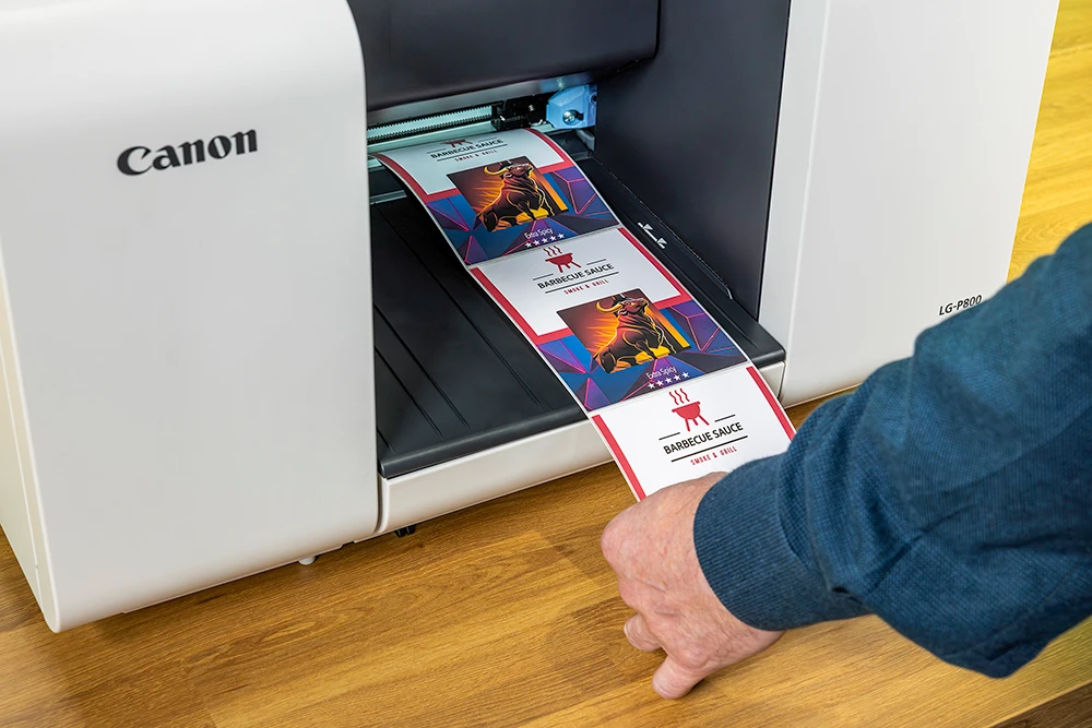 Close up of a Canon LG-P800 label printer feeding out a strip of full colour barbecue sauce labels, each printed with a bull illustration and an Extra Spicy flash. Shown to illustrate the food and beverage use case.