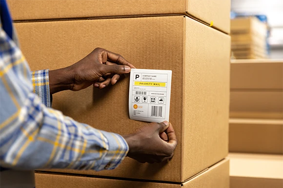 A worker's hands press a freshly printed Priority Mail shipping label, carrying a barcode and an address block, onto a sealed corrugated carton, with more stacked cartons behind. Shown to illustrate the warehouse and shipping use case.