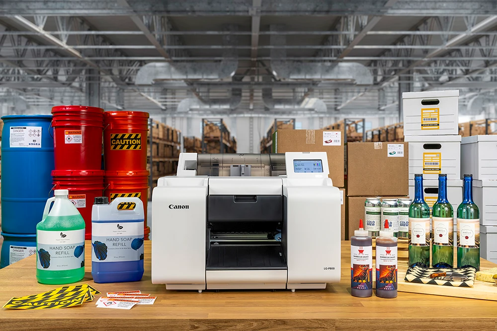 A Canon LG-P800 label printer on a bench inside an industrial facility with steel pallet racking behind it, surrounded by a blue chemical drum and red pails carrying Danger, Warning and Caution hazard labels, plus corrugated cartons and archive boxes with barcode labels. Shown to illustrate the manufacturing use case.