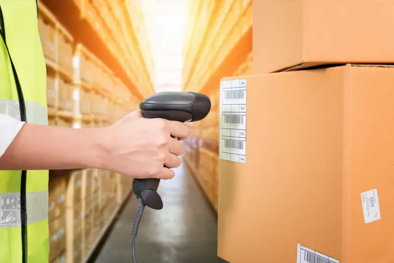 A worker scanning a barcode label on cartons in a warehouse aisle