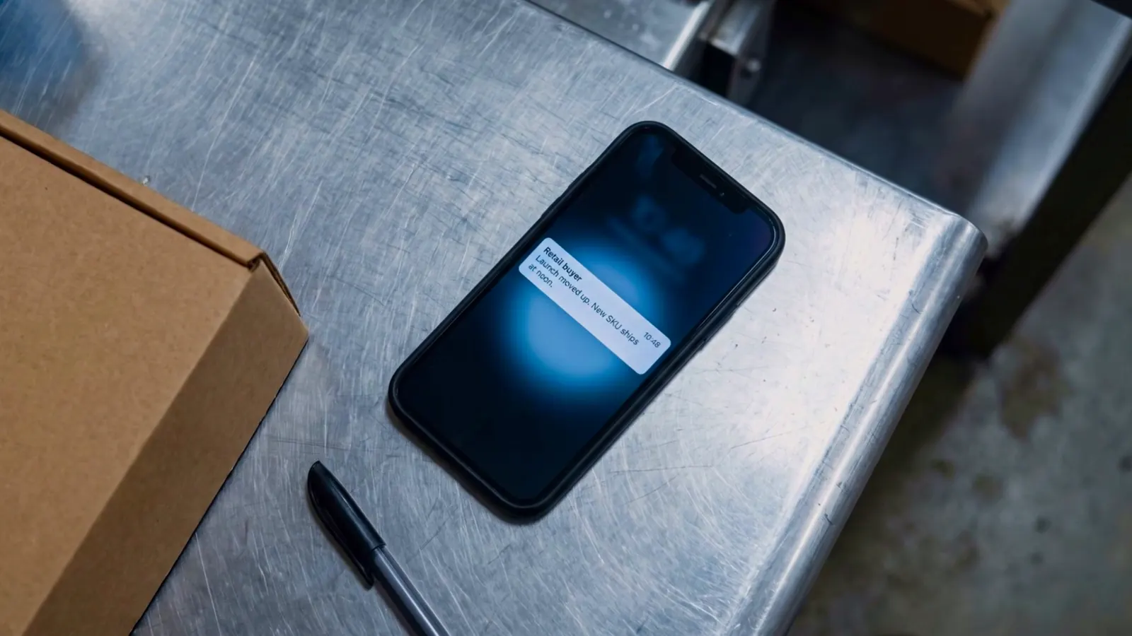 A phone on a steel table shows a message about a new product shipping at noon.