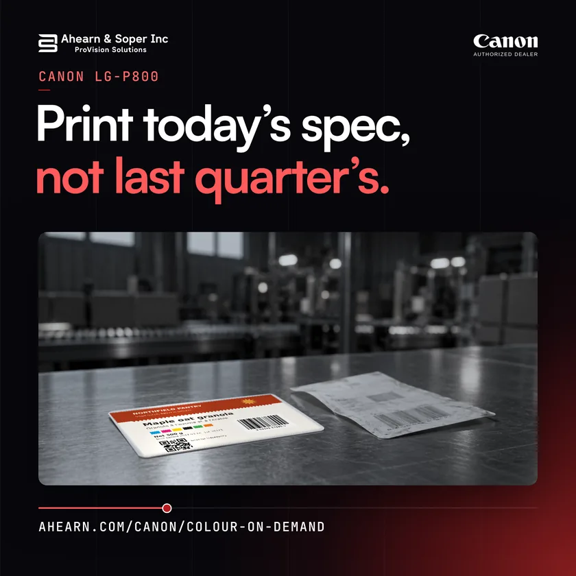 A freshly printed colour label beside an old grey pre-printed label on a steel table in a plant. Text: Print today’s spec, not last quarter’s.