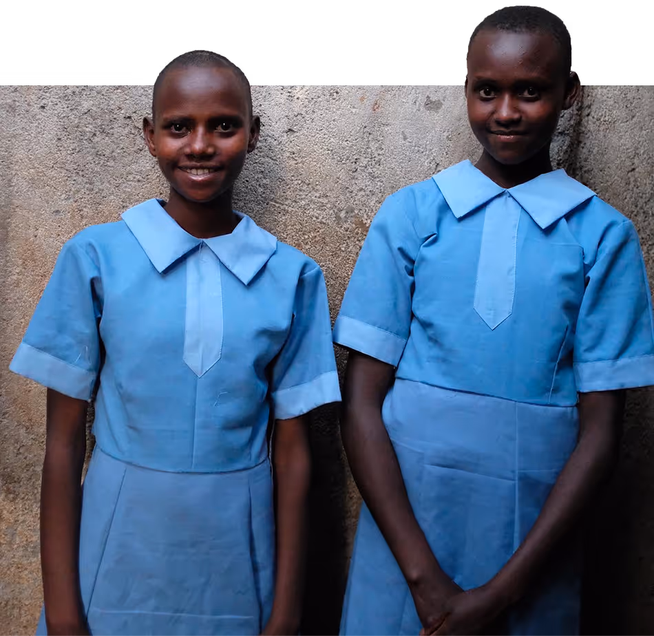 Two smiling Kenyan schoolgirls wearing light blue uniforms standing against a textured wall.