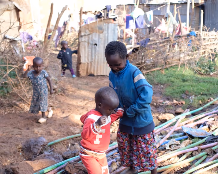 Two young children interacting on a dirt path surrounded by wooden and metal structures, with clothes hanging in the background and other children walking nearby.