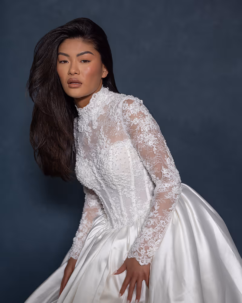 Vrouw met lang zwart haar die een gedetailleerde witte kanten trouwjurk draagt met parelversieringen en een satijnen rok, poseert voor een donkere achtergrond.