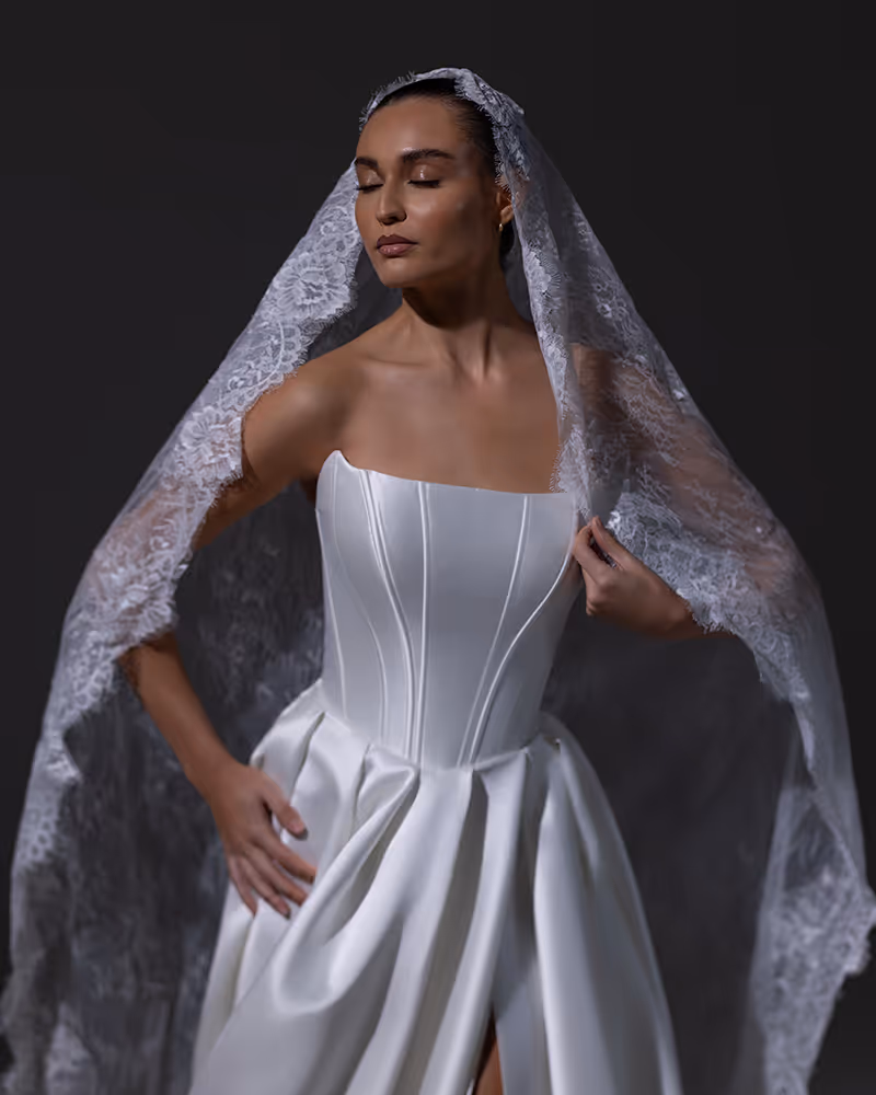 Vrouw in witte strapless trouwjurk met kanten sluier die over haar hoofd hangt en gesloten ogen heeft.