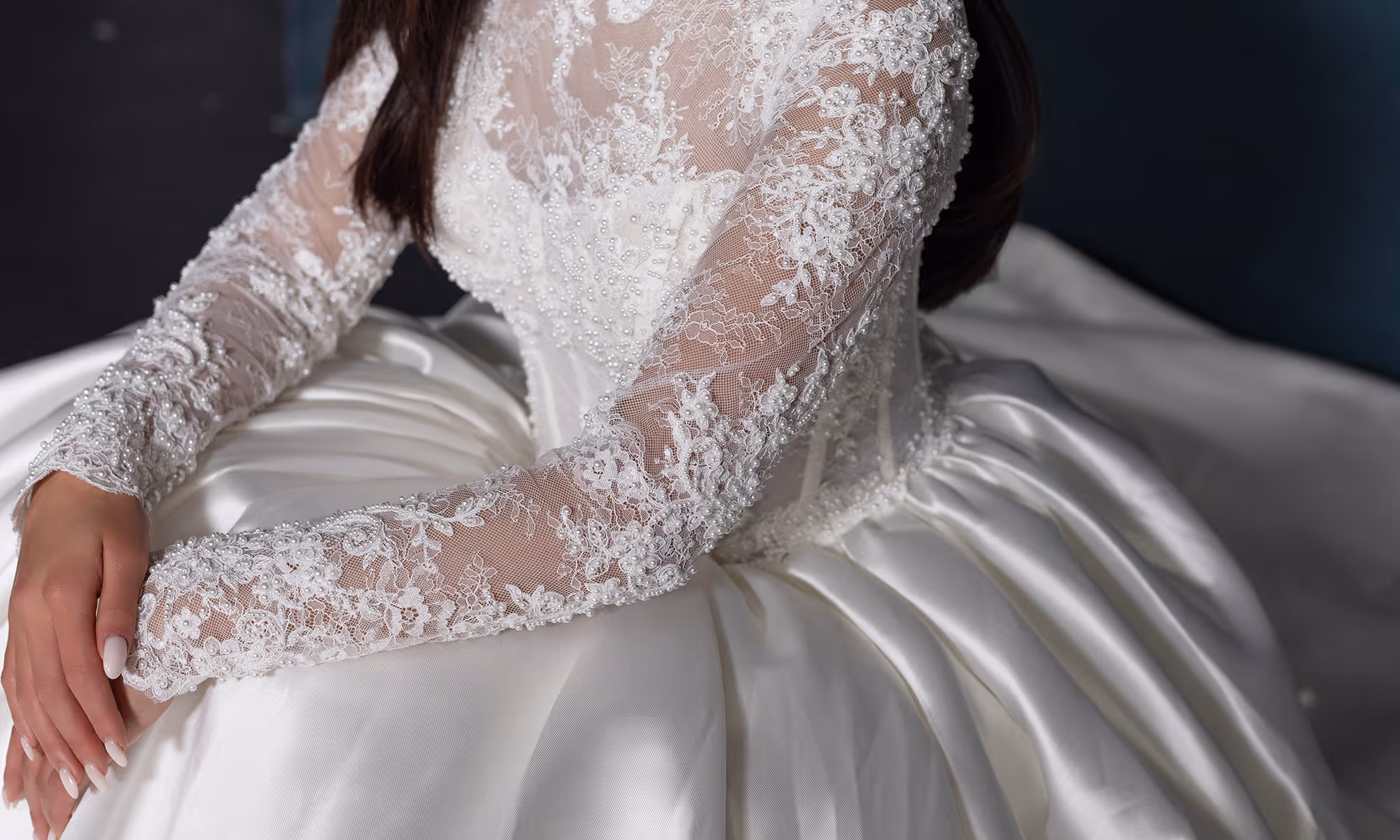 Close-up van een bruid in een witte trouwjurk met kanten mouwen en pareldetails, zittend met gevouwen handen.