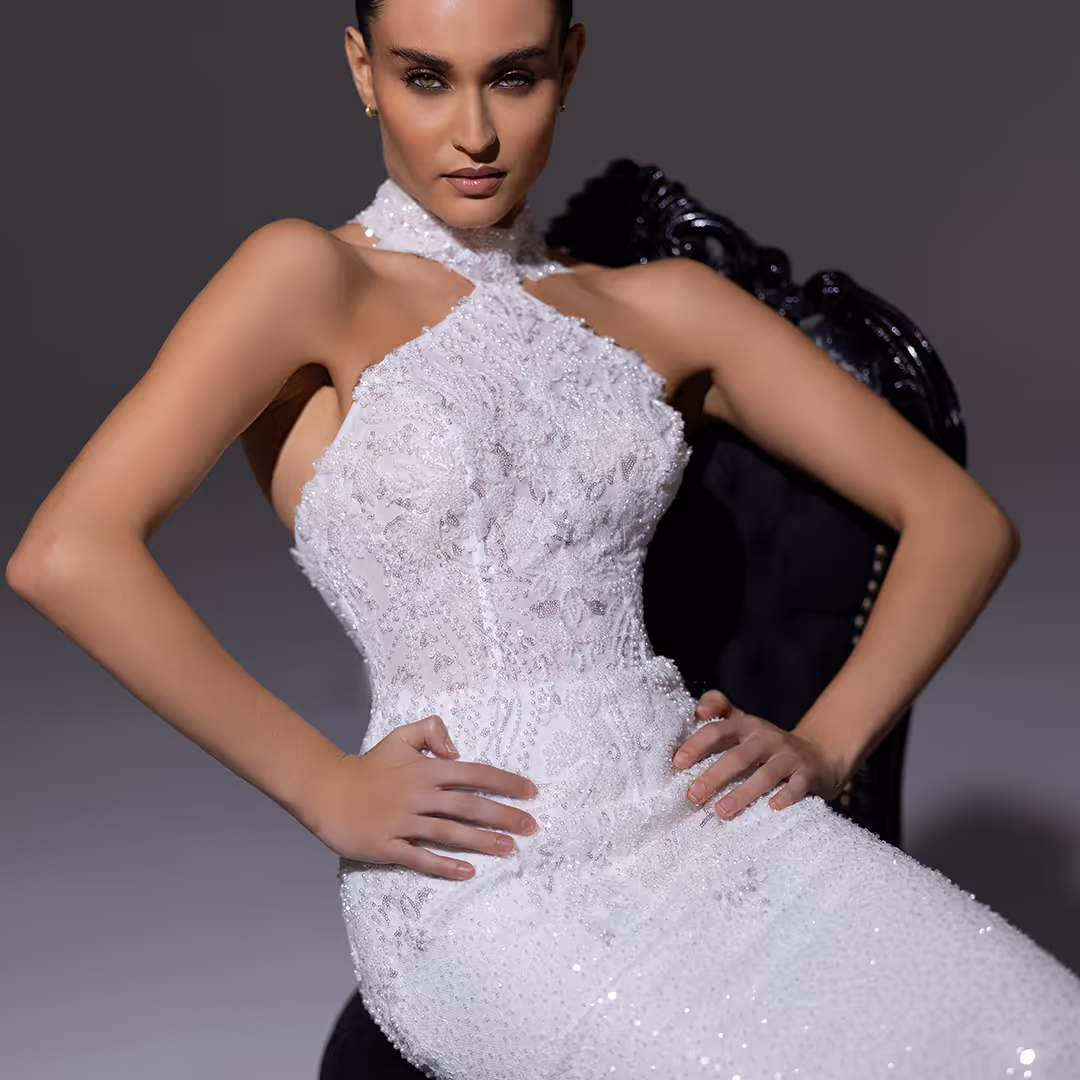 Model draagt een witte, geborduurde avondjurk zittend op een zwart sierstoel met handen in haar taille.