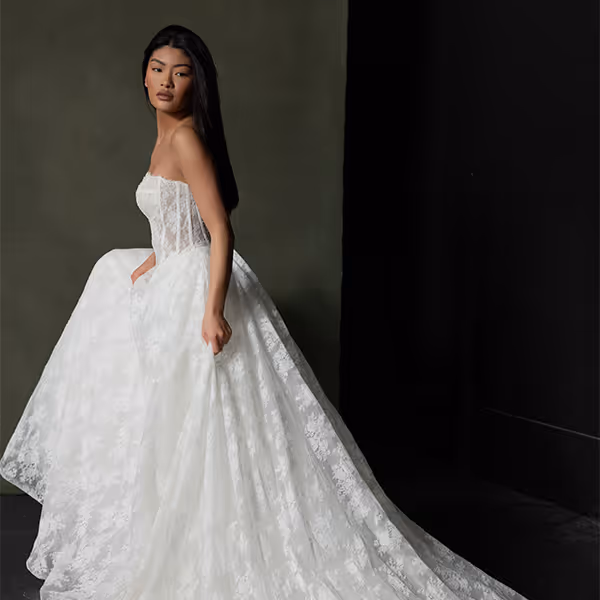 Vrouw in een strapless trouwjurk met een lange, kanten sleep tegen een effen donkere achtergrond.