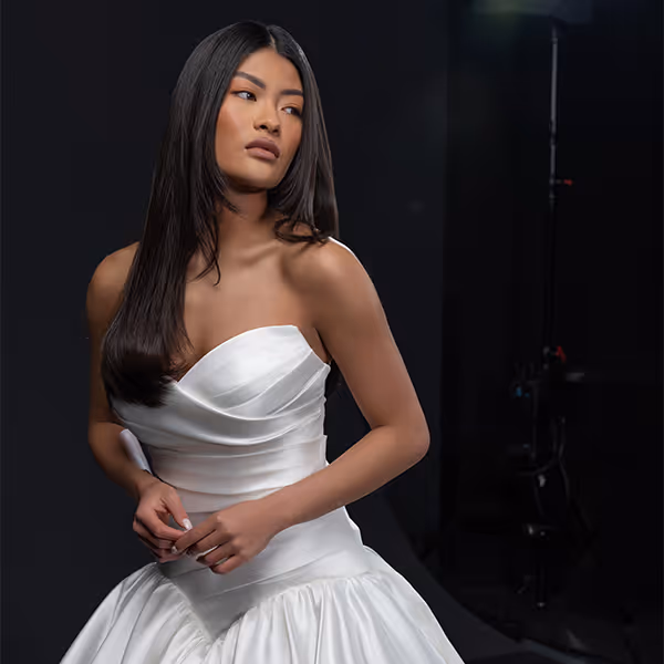 Vrouw met lang zwart haar in een strapless witte trouwjurk, poserend tegen een donkere achtergrond.