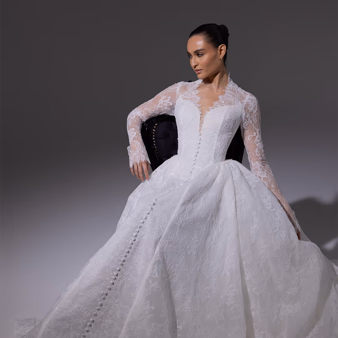 Vrouw in een kanten witte trouwjurk met lange mouwen en knopen langs het midden, zittend op een donkergekleurde stoel.