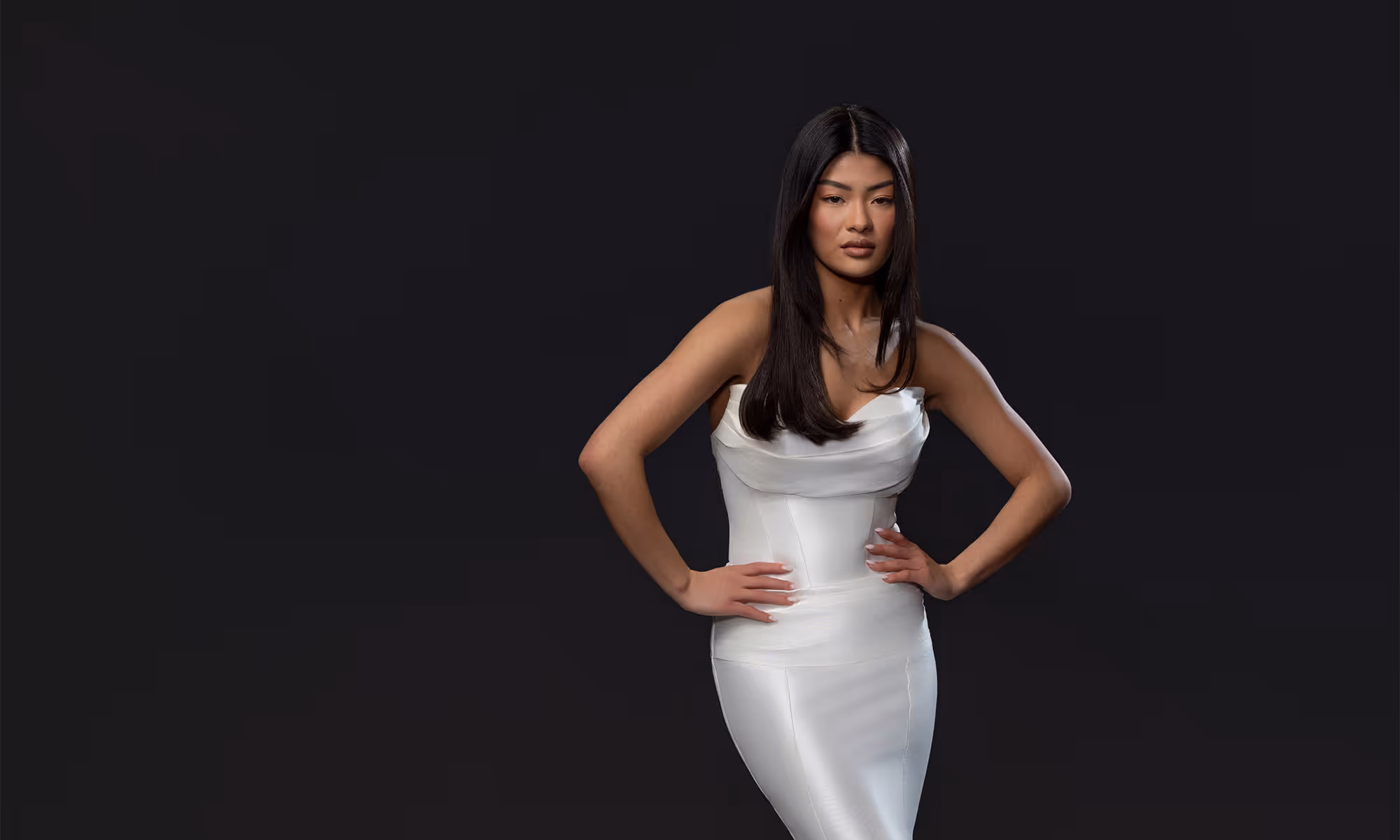 Vrouw met lang zwart haar en witte strapless jurk poseert tegen donkere achtergrond met handen in haar zij.