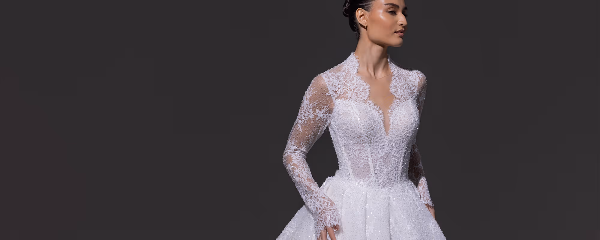 Vrouw met een kanten witte trouwjurk met lange mouwen, staand tegen een donkere achtergrond.