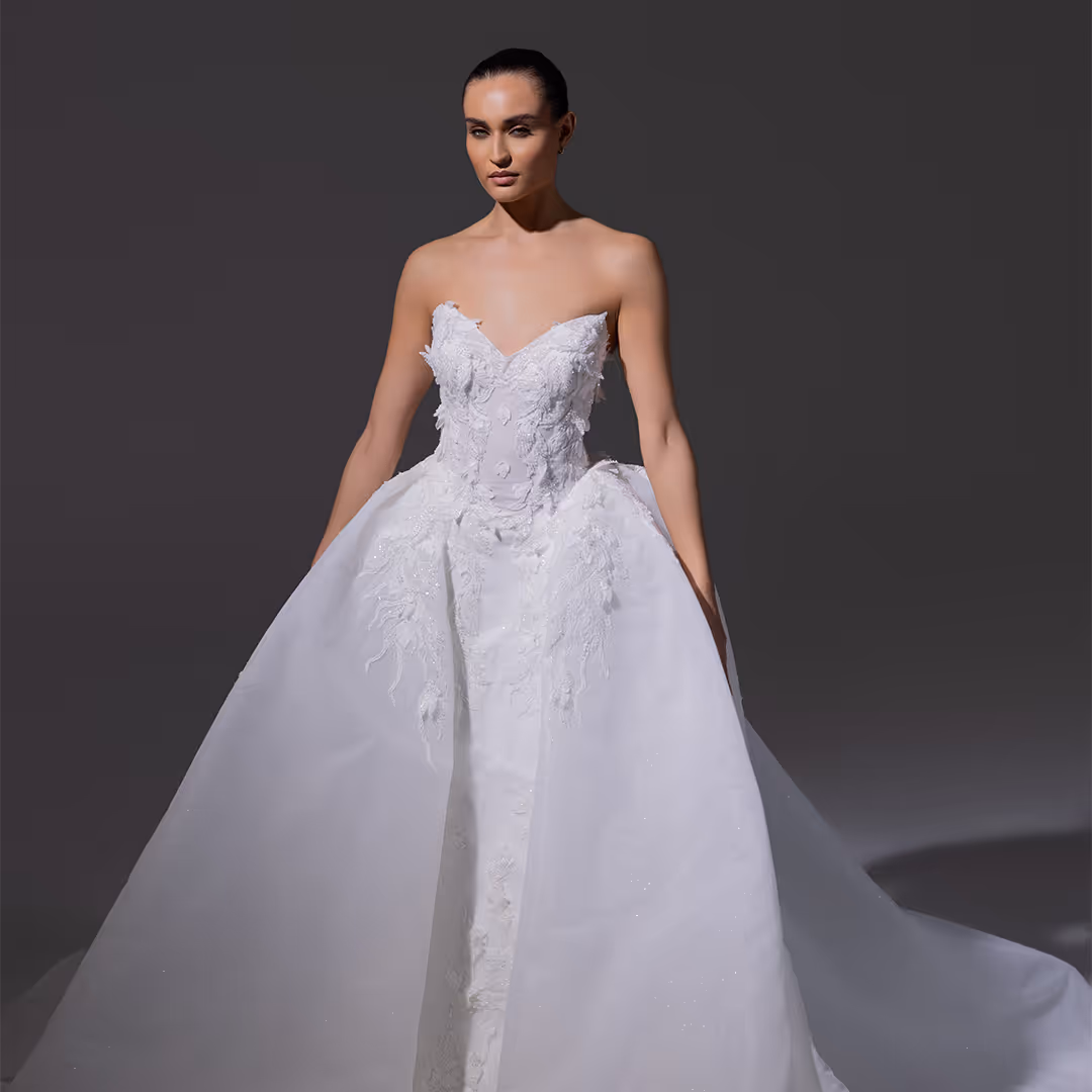 Vrouwelijk model met een strapless witte bruidsjurk met borduursels en een wijde rok, staand tegen een donkere achtergrond.