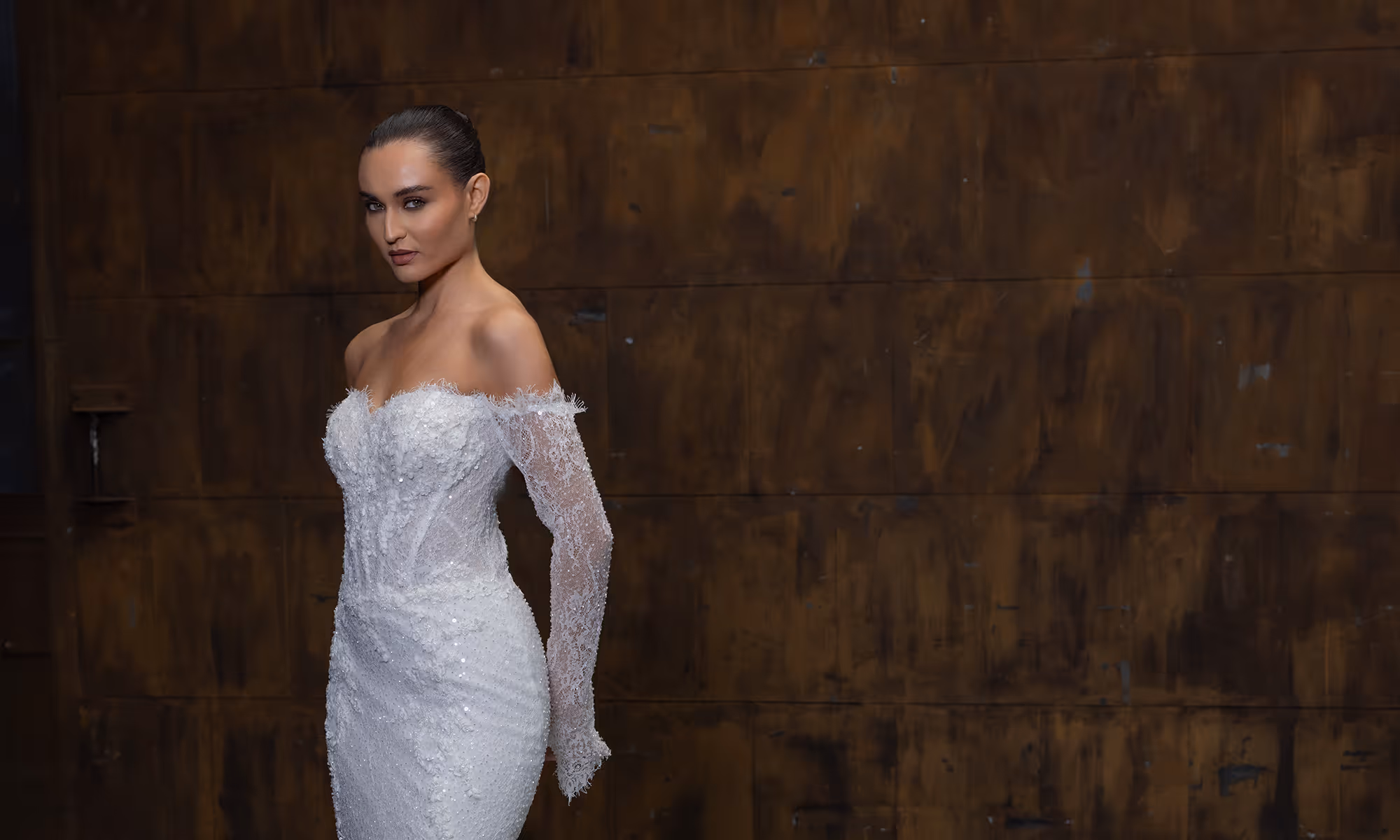 Vrouw in een elegante witte kanten off-shoulder trouwjurk met één lange mouw, staand tegen een donkere houten achtergrond.