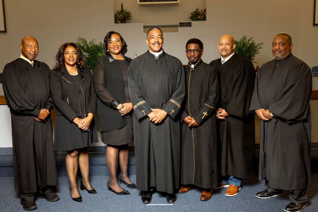 Seven people standing indoors in formal attire, five men wearing black robes with gold cross details and two women in black dresses and blazers.