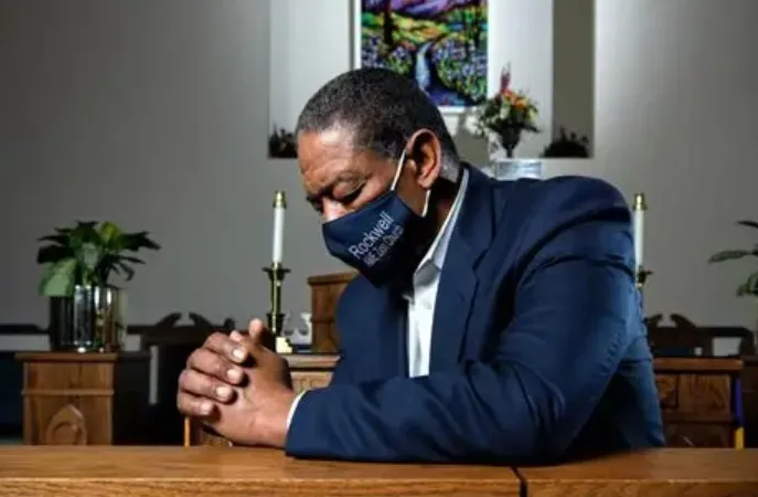 Man wearing a face mask with 'Rockwell' printed on it, praying with clasped hands at a church pew.