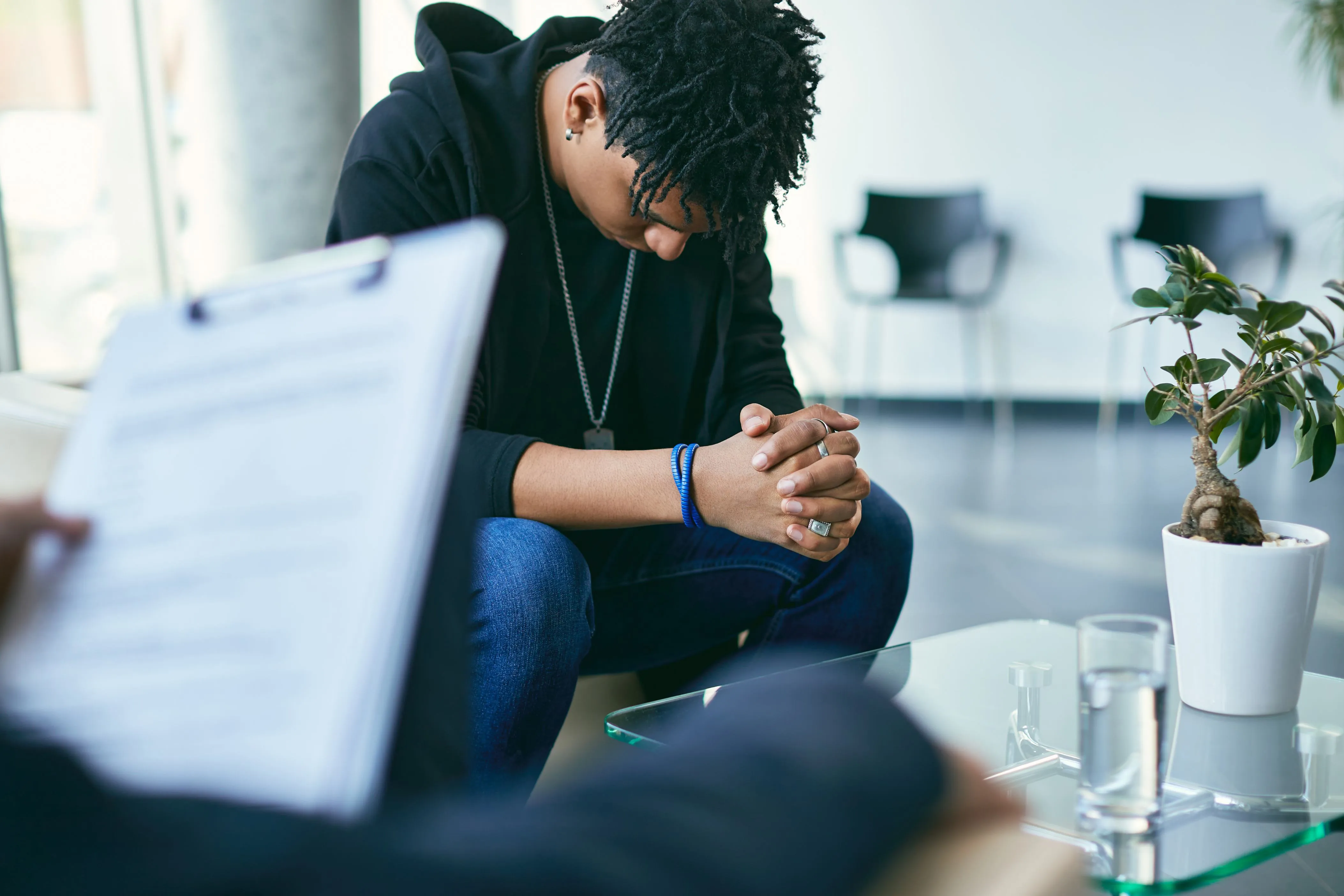 Young man with clasped hands and head bowed during a therapy session in a modern office.