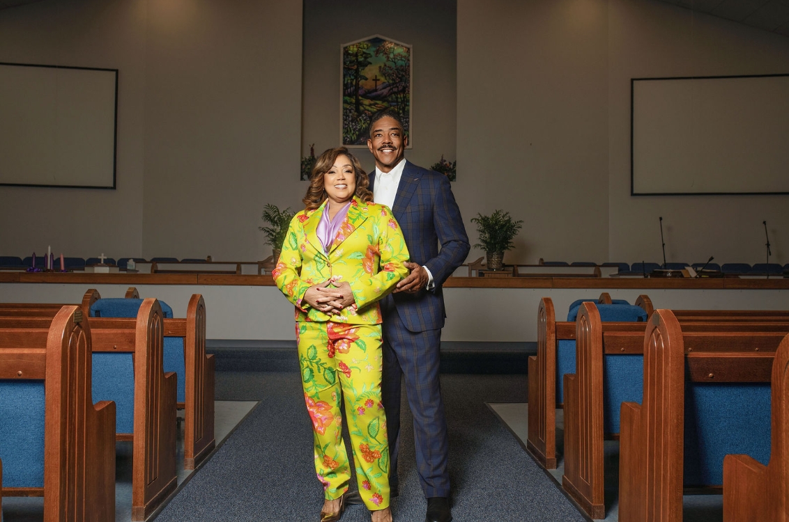 A man in a blue suit and a woman in a bright yellow floral suit smiling and standing together in the aisle of a church.