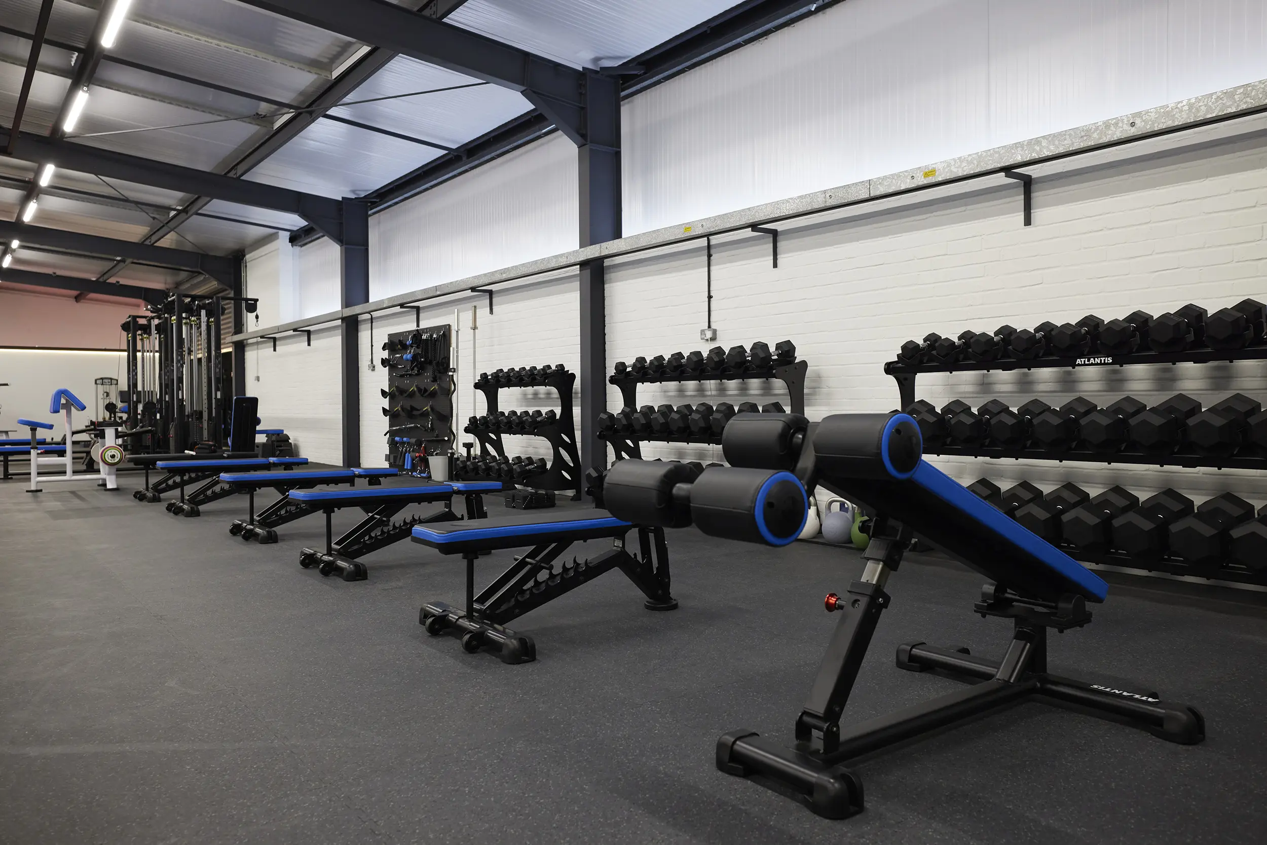 Modern gym interior with adjustable weight benches, dumbbell racks, and kettlebells along a white brick wall.