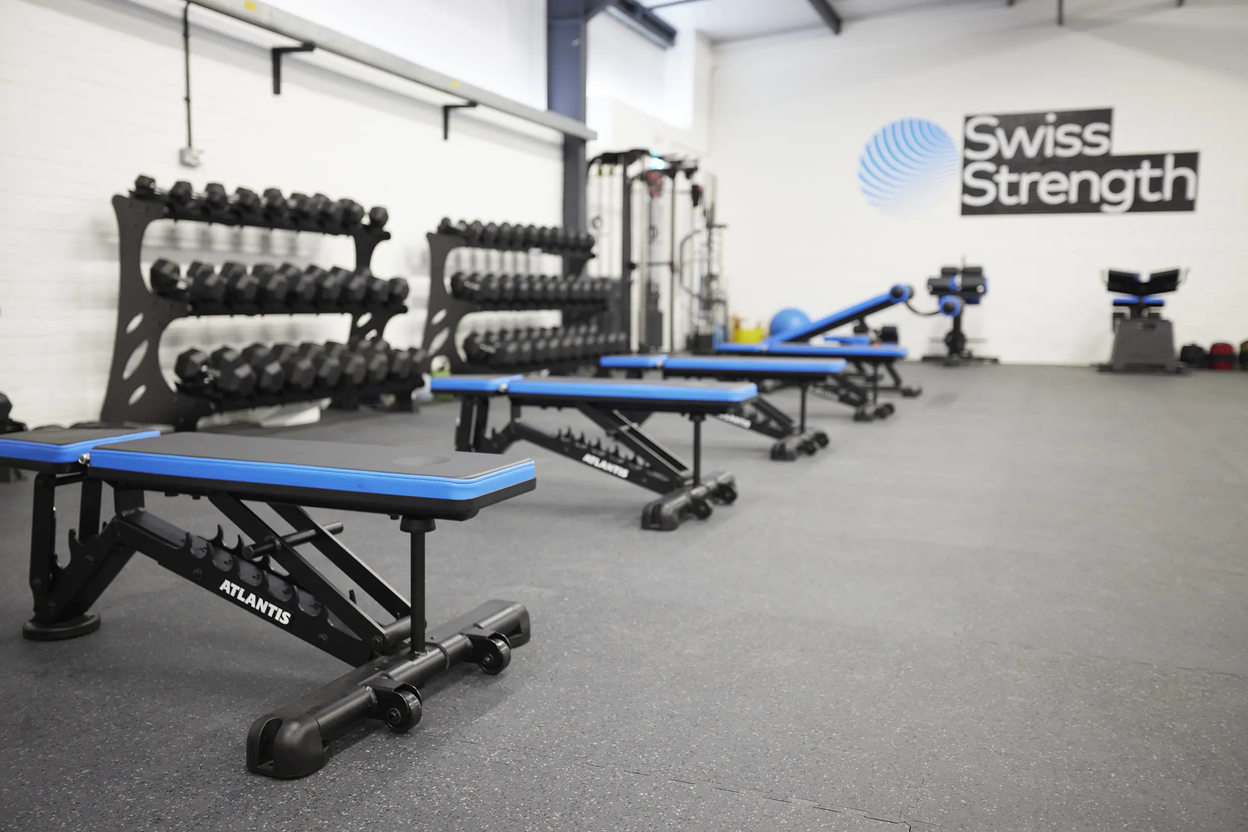 Empty gym with rows of blue workout benches and racks of dumbbells under Swiss Strength signage.