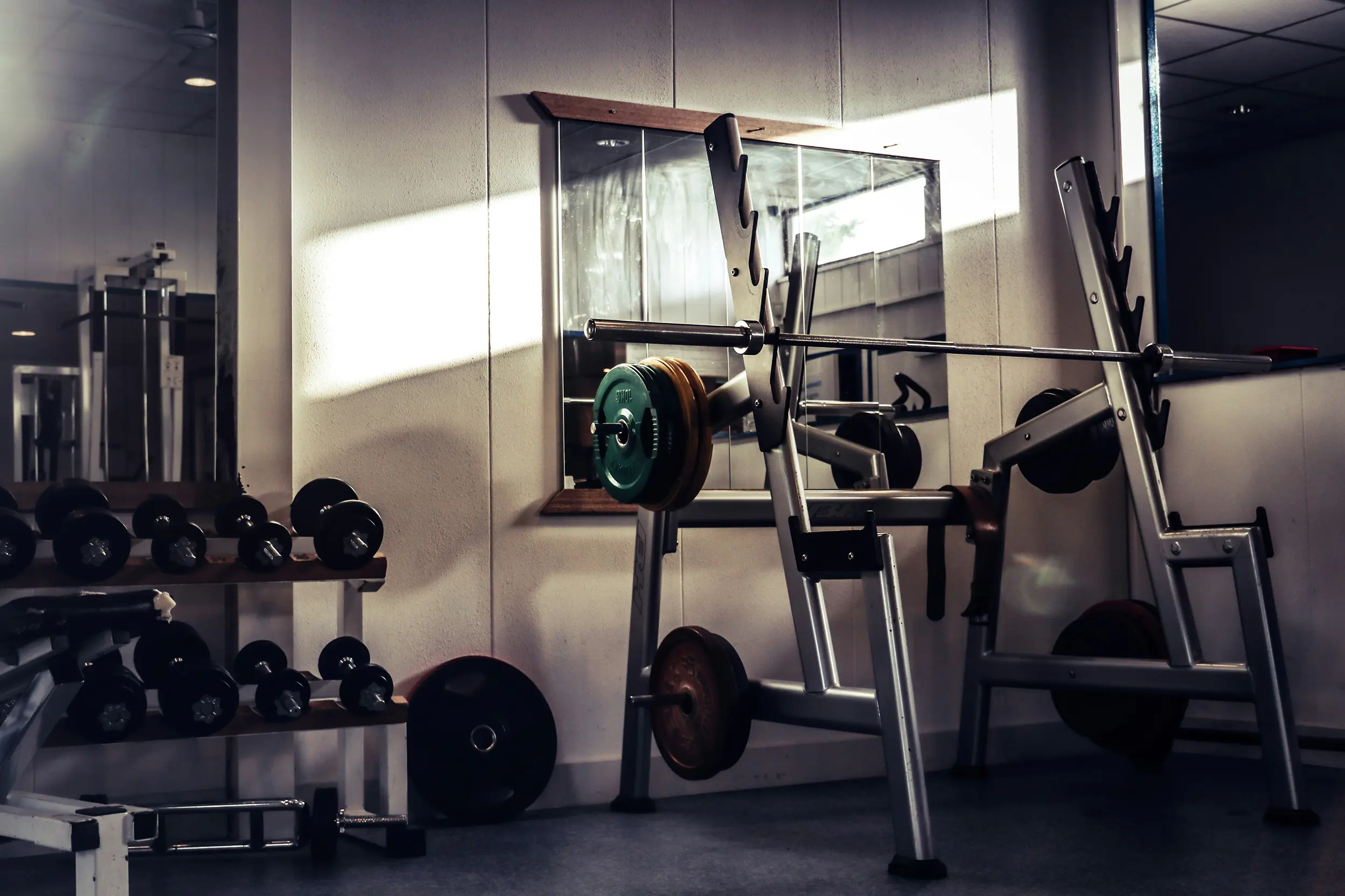 Weightlifting bench with barbell and weight plates in a gym with dumbbells on racks nearby.