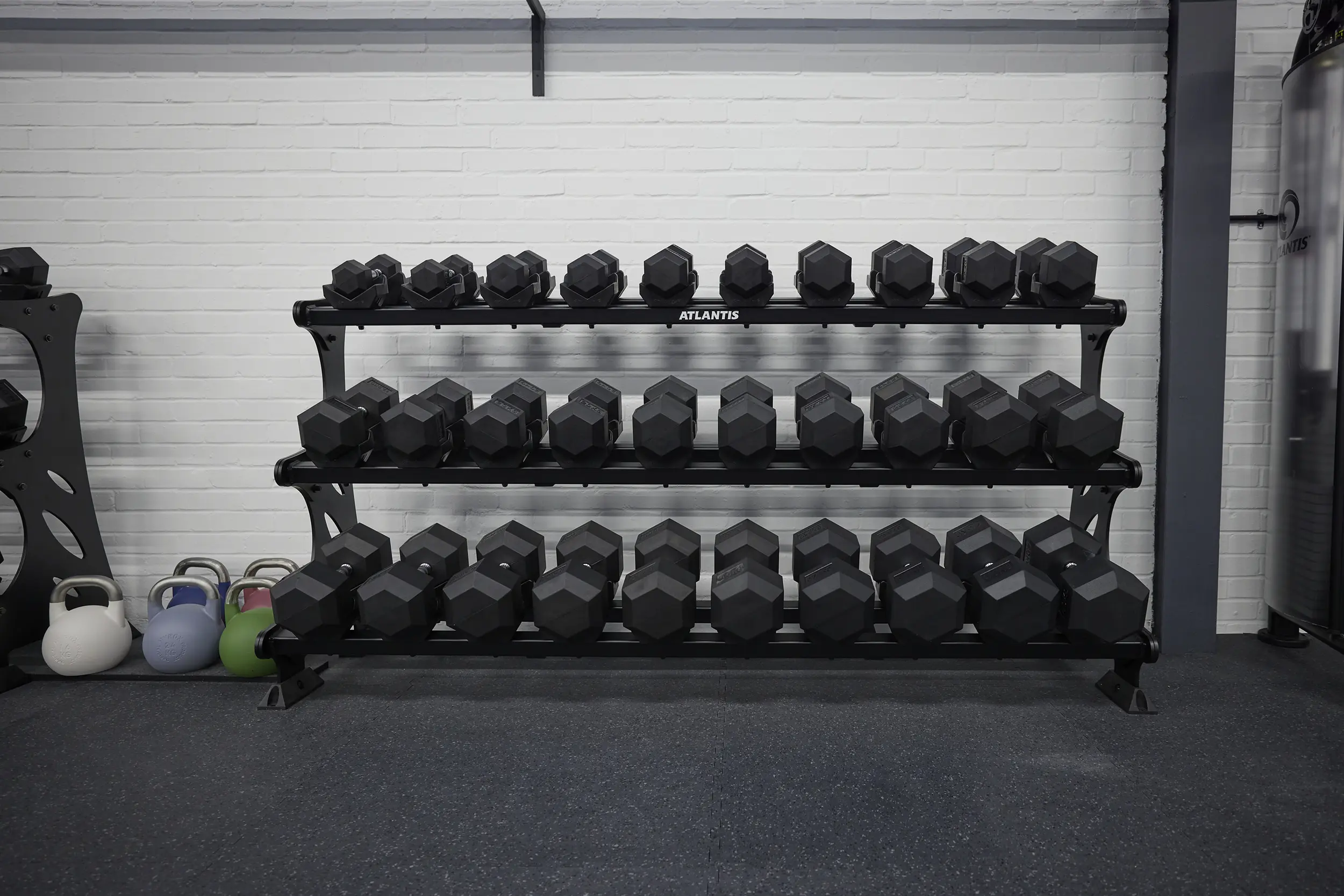 Three-tier rack filled with various sizes of black hexagonal dumbbells and colorful kettlebells on the floor to the left in a gym.