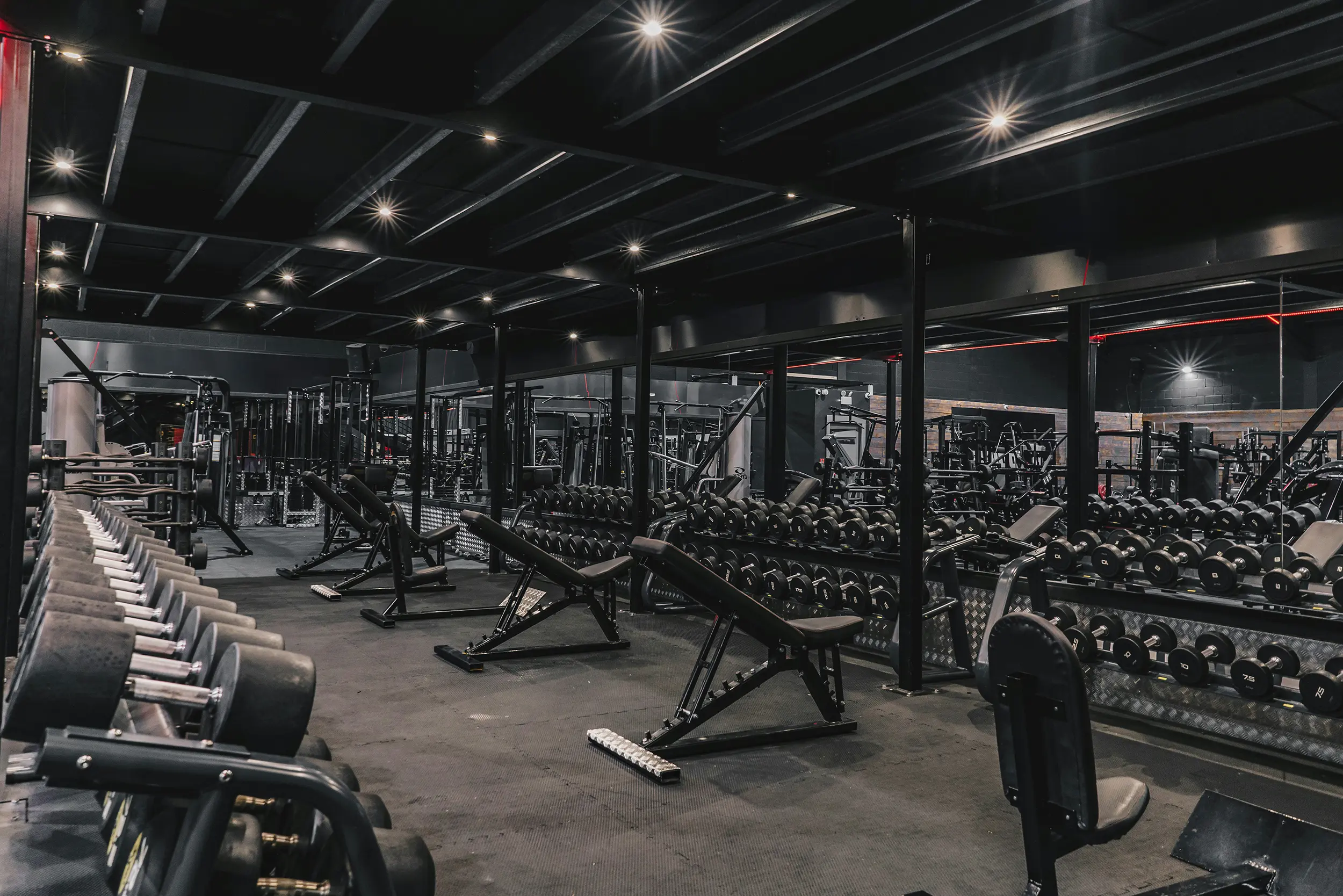Modern gym interior with rows of dumbbells, adjustable benches, and mirrored walls under black ceiling lights.
