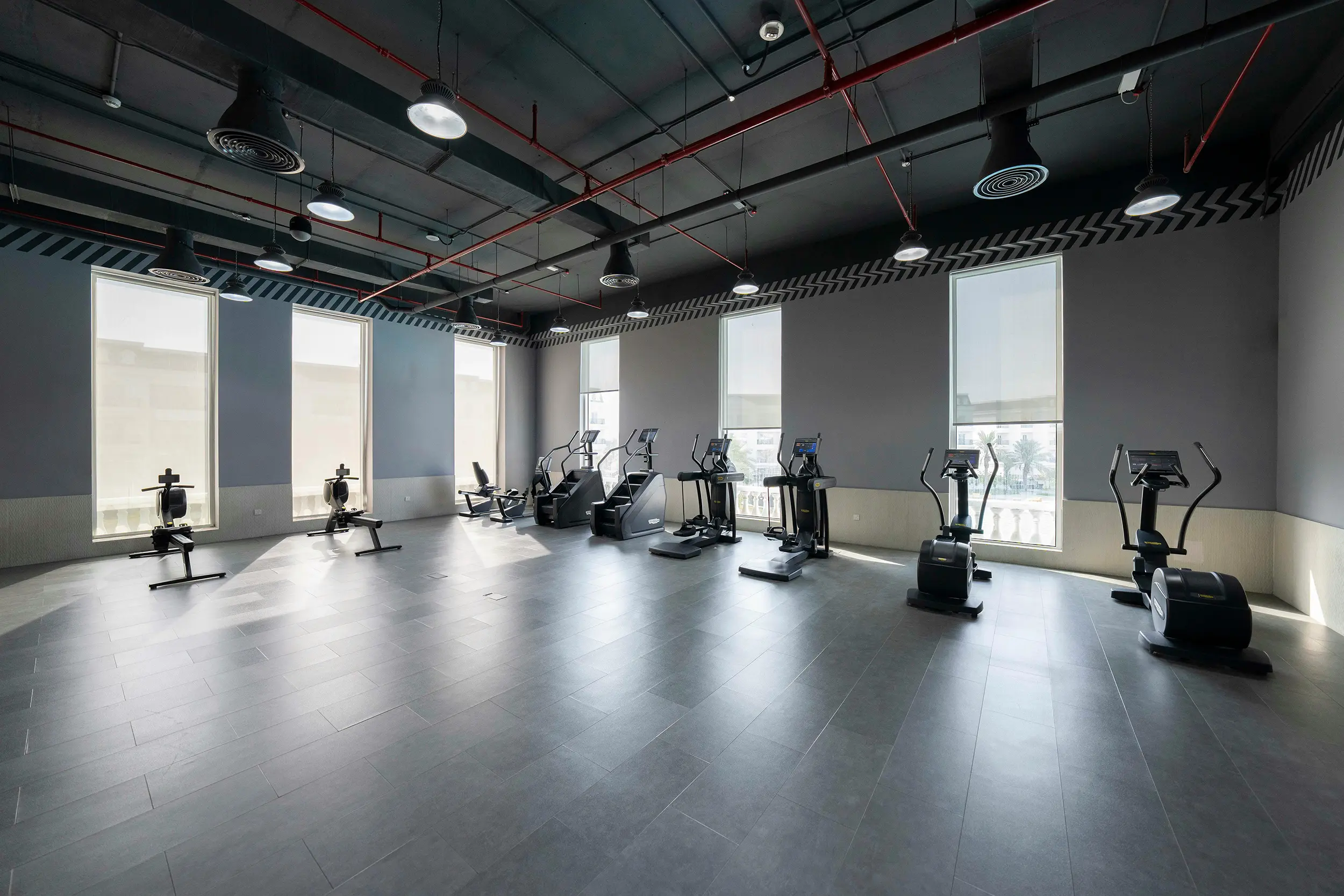 Spacious gym with exercise machines including rowing machines, elliptical trainers, and stationary bikes lined up against large windows.