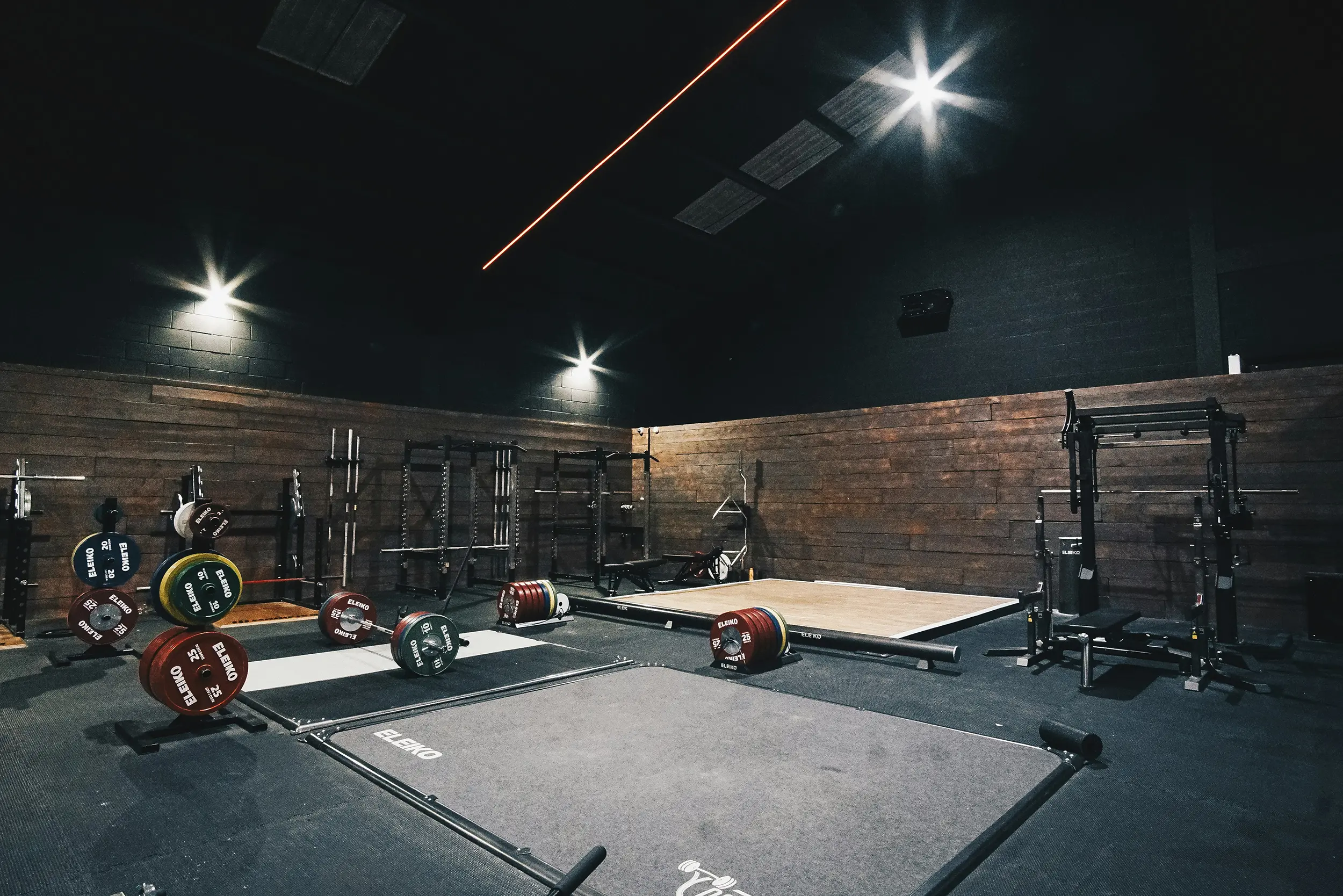 Modern gym interior with weightlifting platforms, barbells loaded with colorful weight plates, and black squat racks against wooden paneled walls.