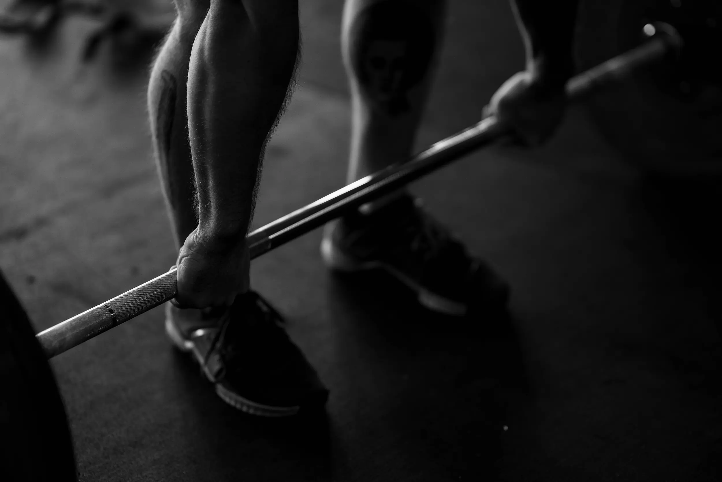 Close-up of a person gripping a barbell in a gym, focusing on forearms and legs with athletic shoes.