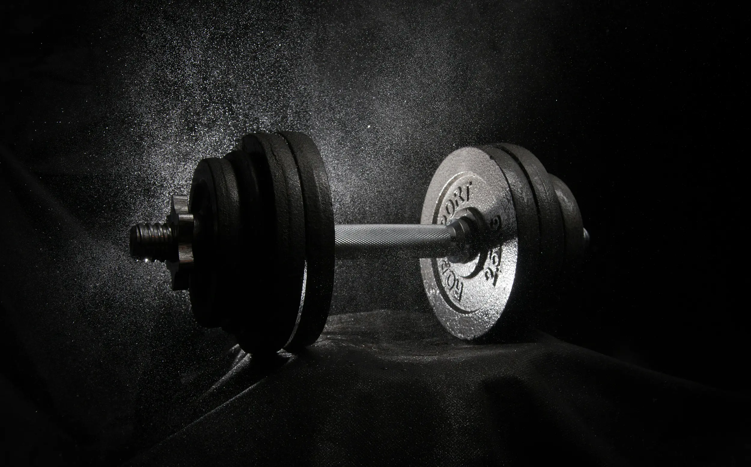 Black and silver adjustable dumbbell with powder particles floating around it on a black background.