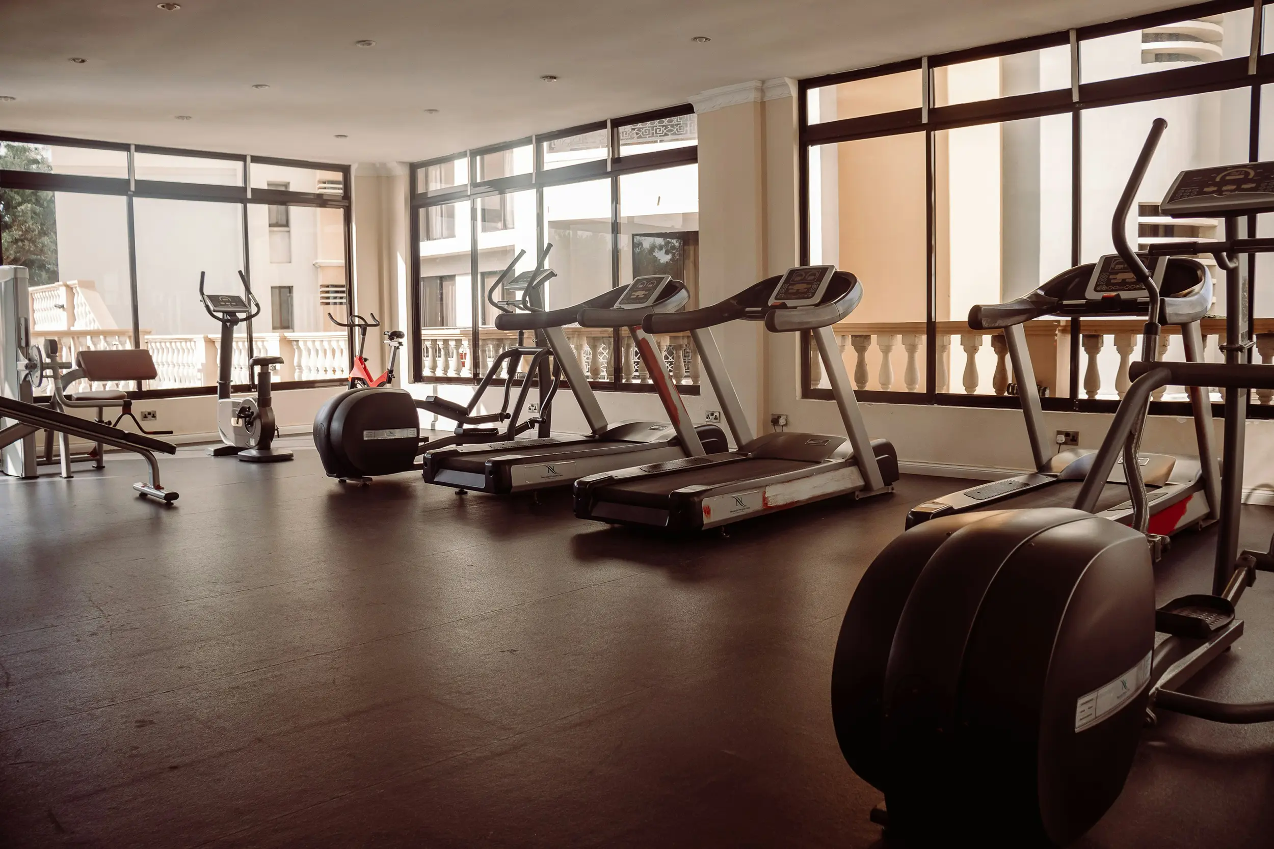 Empty gym room with treadmills, exercise bikes, and elliptical machines near large windows.