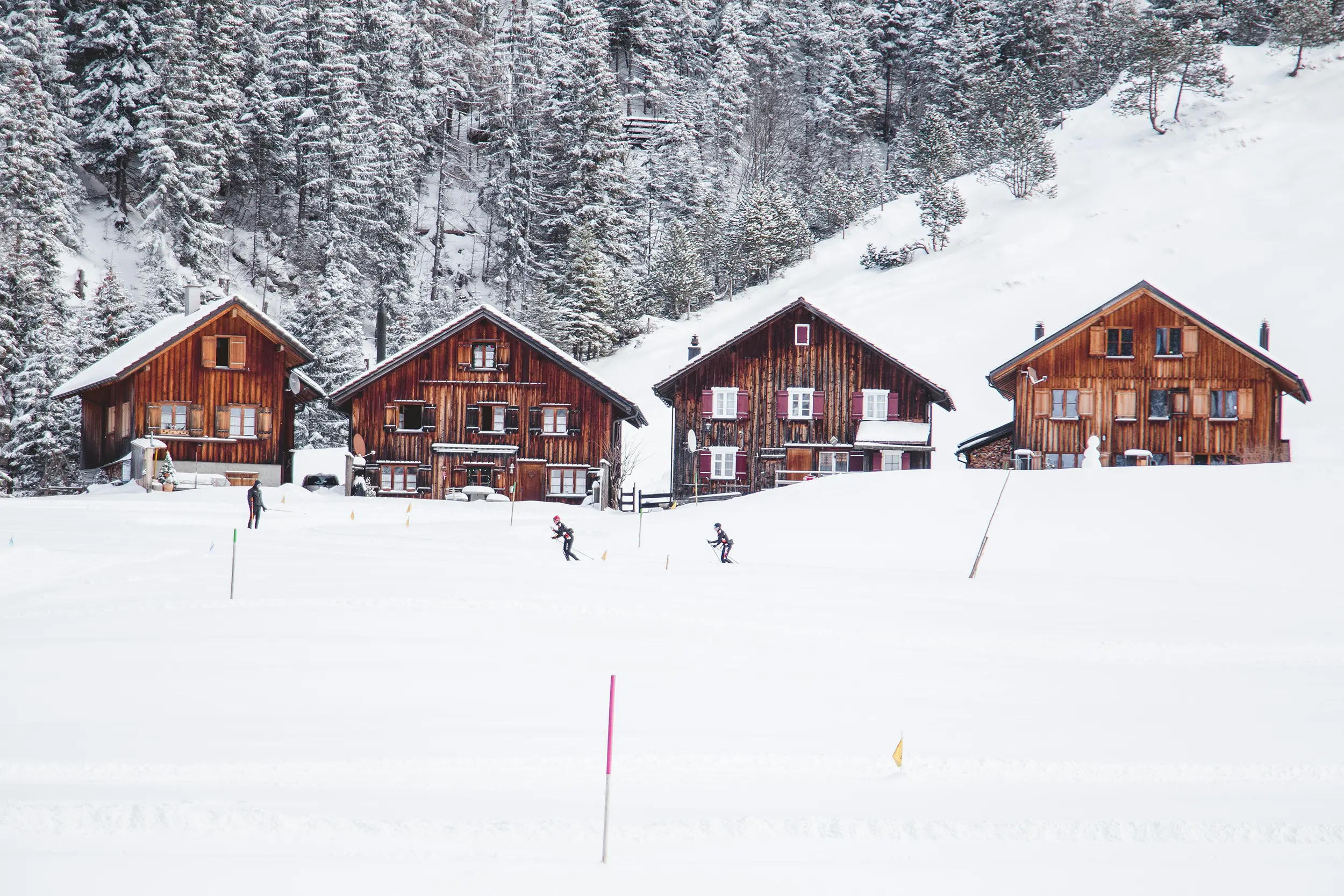 Four wooden cabins in a snowy landscape with three people skiing on a snow-covered field in front.
