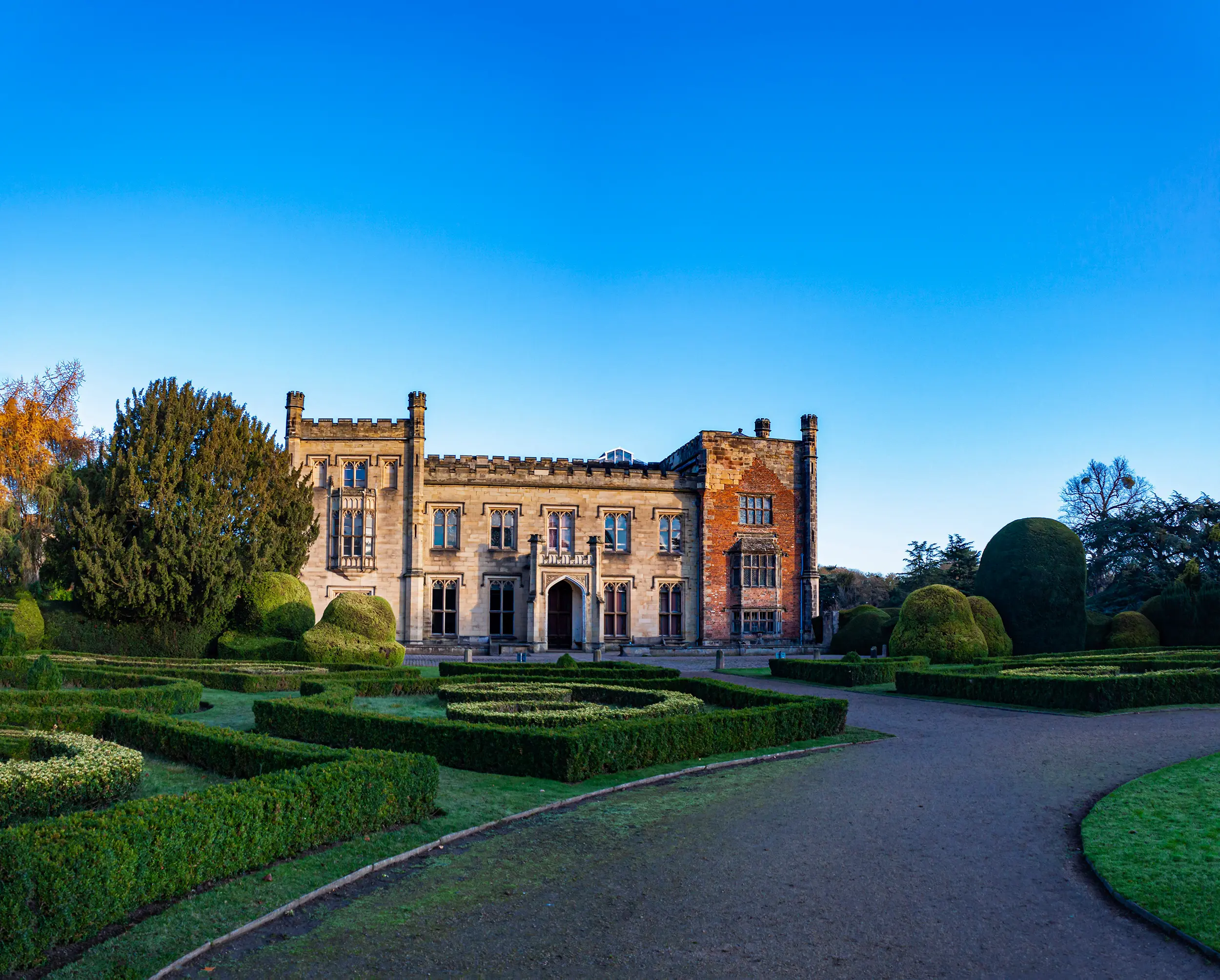 Historic stone manor with battlements surrounded by manicured gardens and clear blue sky.