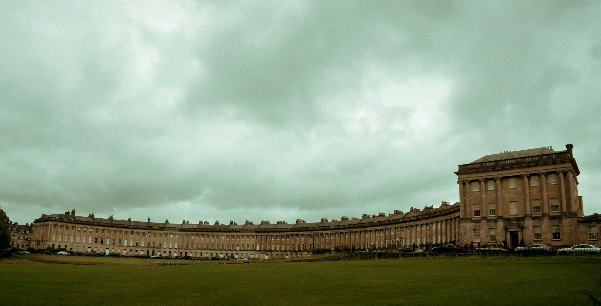 Wide view of the Royal Crescent, a curved row of 18th-century Georgian townhouses in Bath, England, under a cloudy sky.