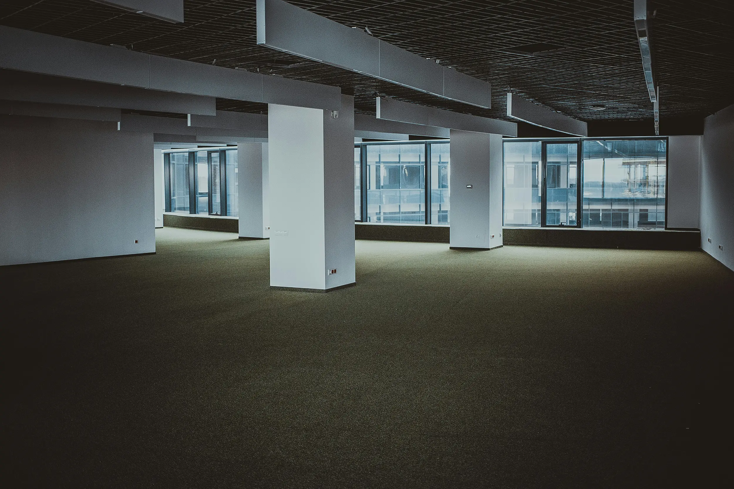 Spacious empty office space with large windows, white columns, and dark carpeted floor.