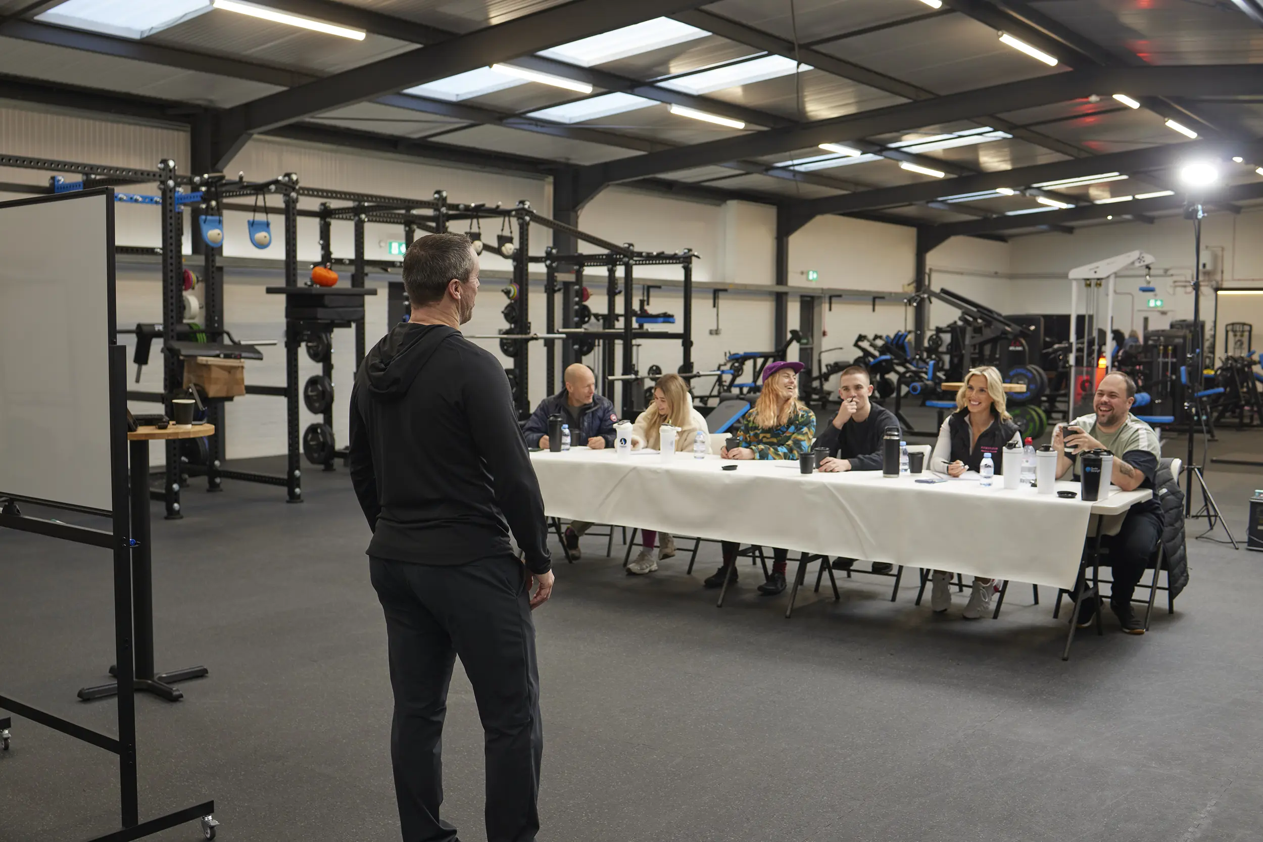Man in black facing a panel of six people seated behind a table in a gym setting with weightlifting equipment.
