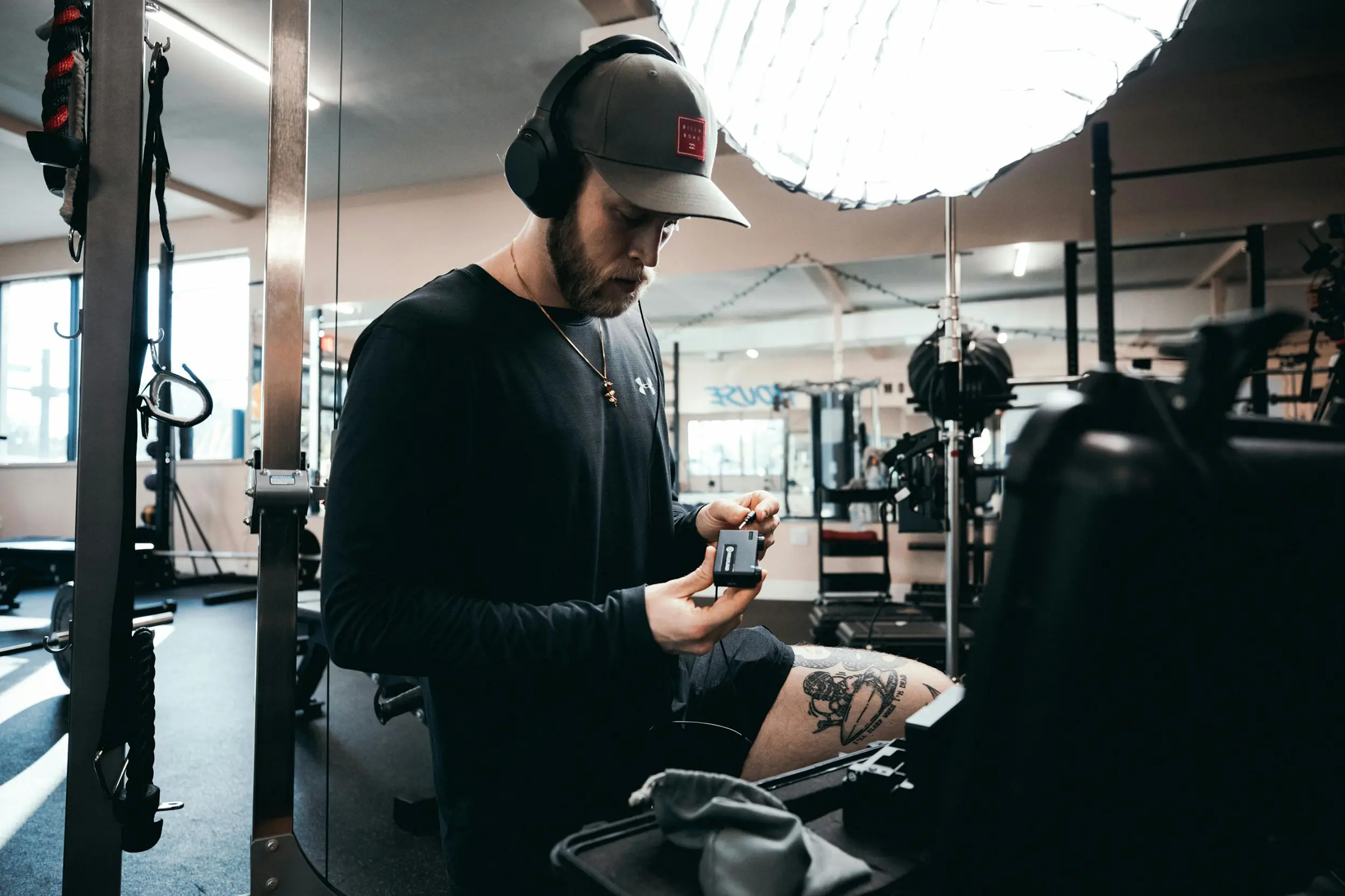 Man wearing headphones and a cap adjusting audio equipment in a gym setting with workout machines in the background.