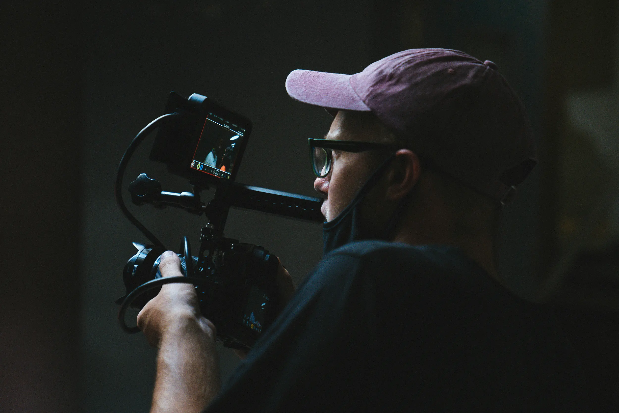 Man wearing glasses and a cap operating a professional camera with an external monitor in a dimly lit setting.