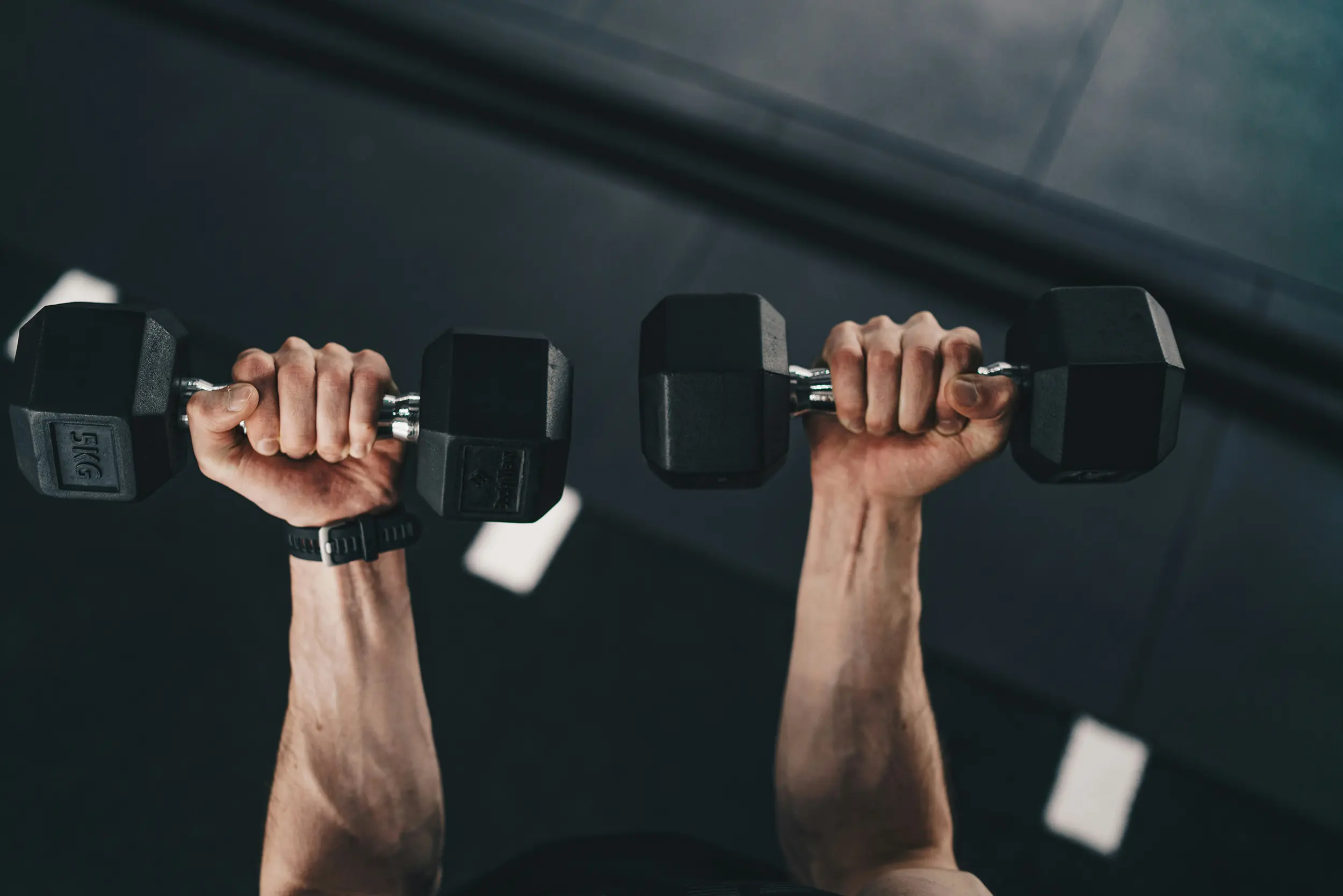 Person lifting two 5kg black hex dumbbells in a gym.
