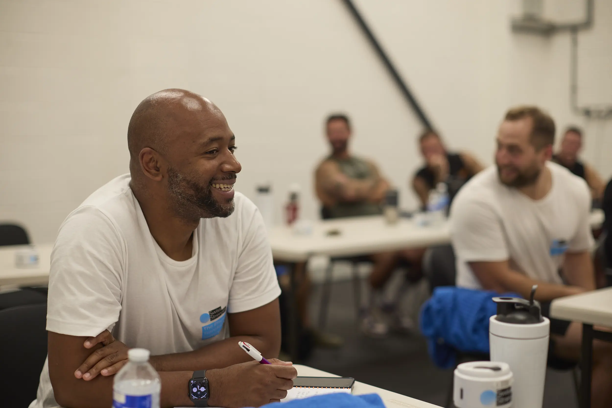 Smiling man in a white t-shirt sitting at a table with a notebook and pen during a group discussion or lecture.