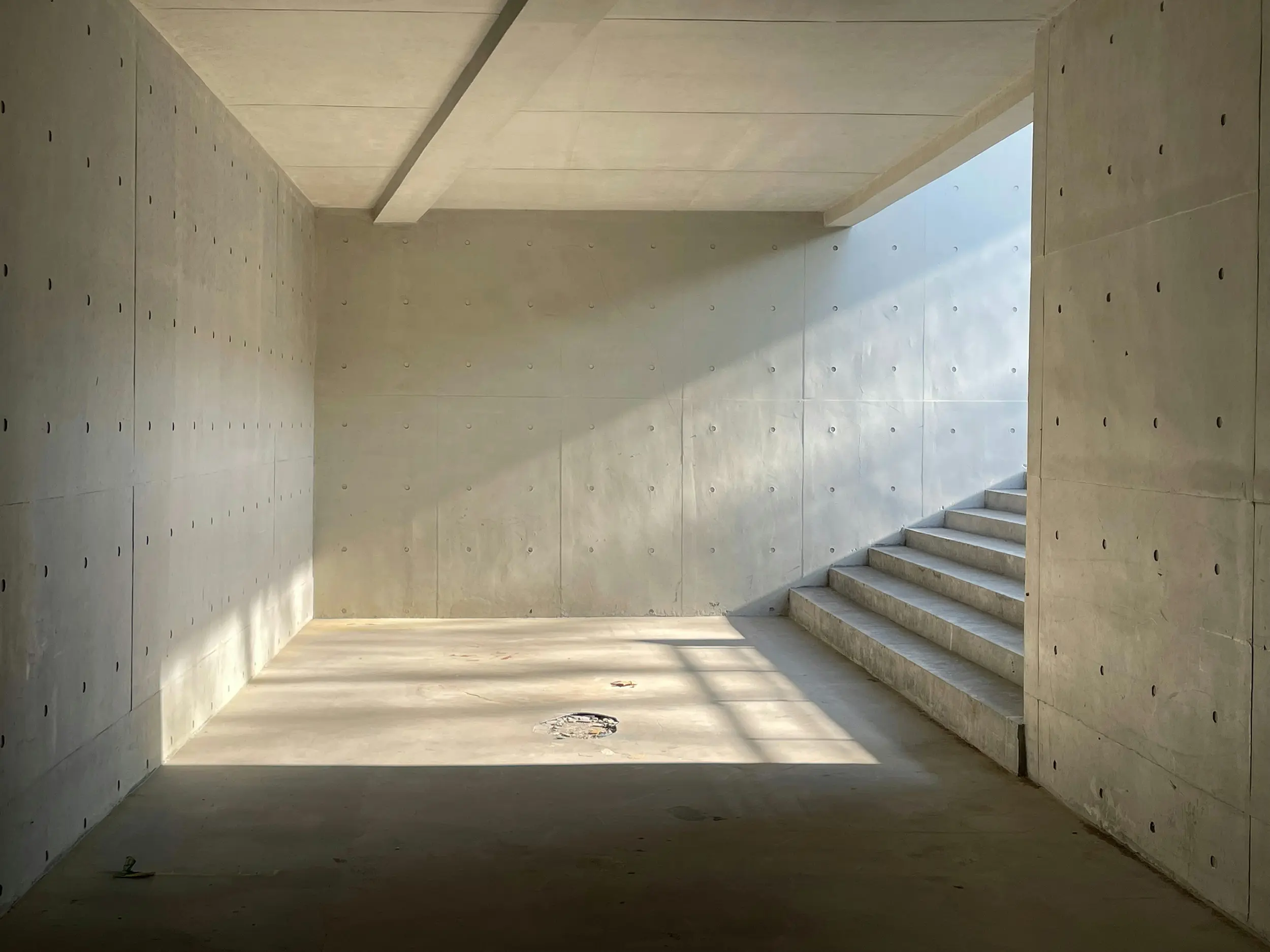 Empty concrete room with stairs on the right and sunlight casting shadows on the floor and wall.