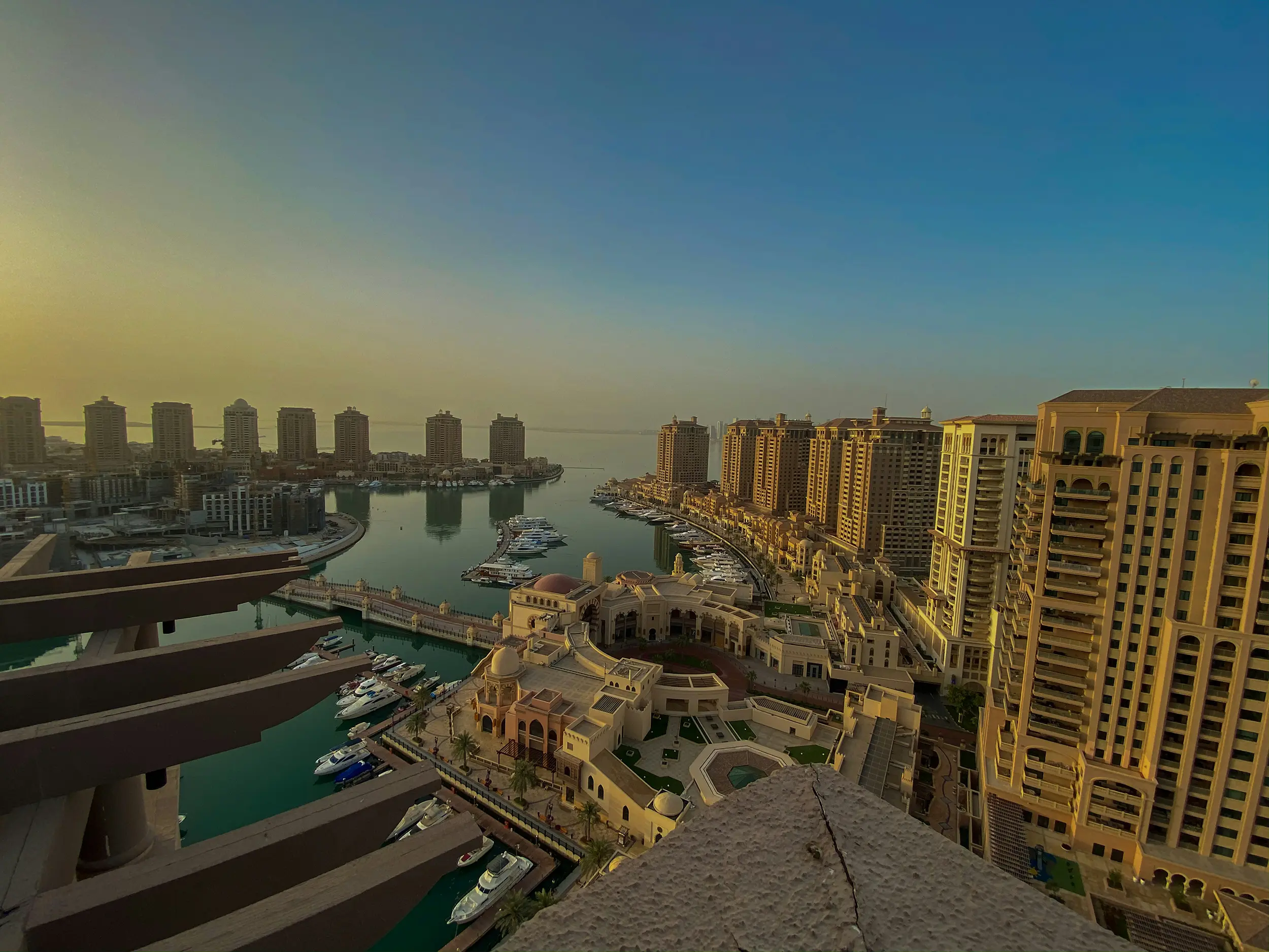 Aerial view of a marina lined with yachts and surrounded by tall residential buildings under a clear sky at sunset.
