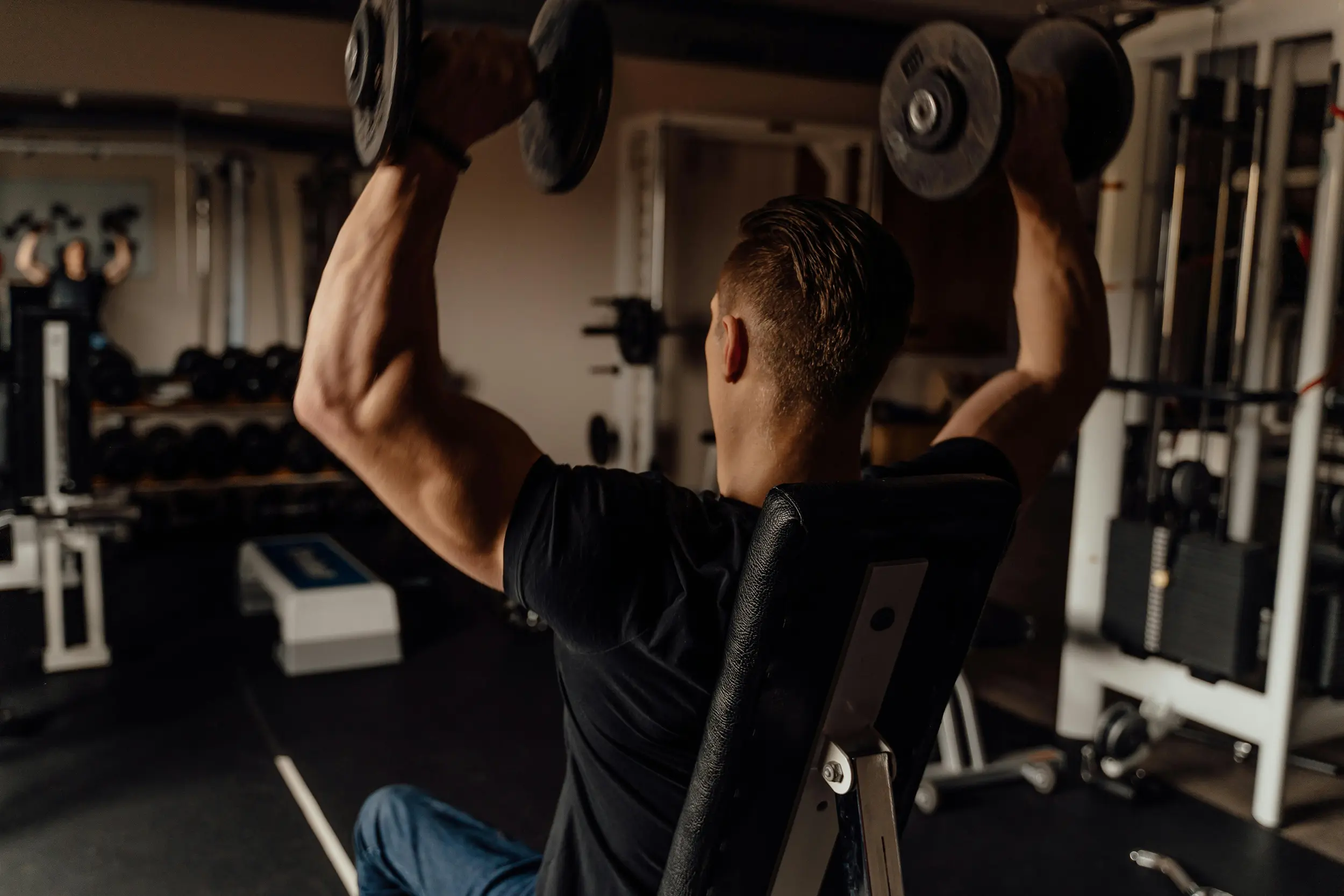 Muscular man in black shirt lifting dumbbells overhead while seated in a gym.