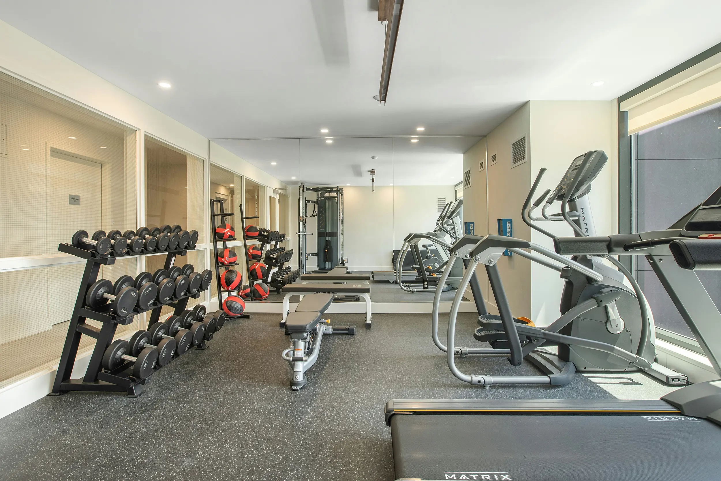 Modern gym room with dumbbells, weight bench, medicine balls, elliptical machines, and treadmill near a large mirror and window.