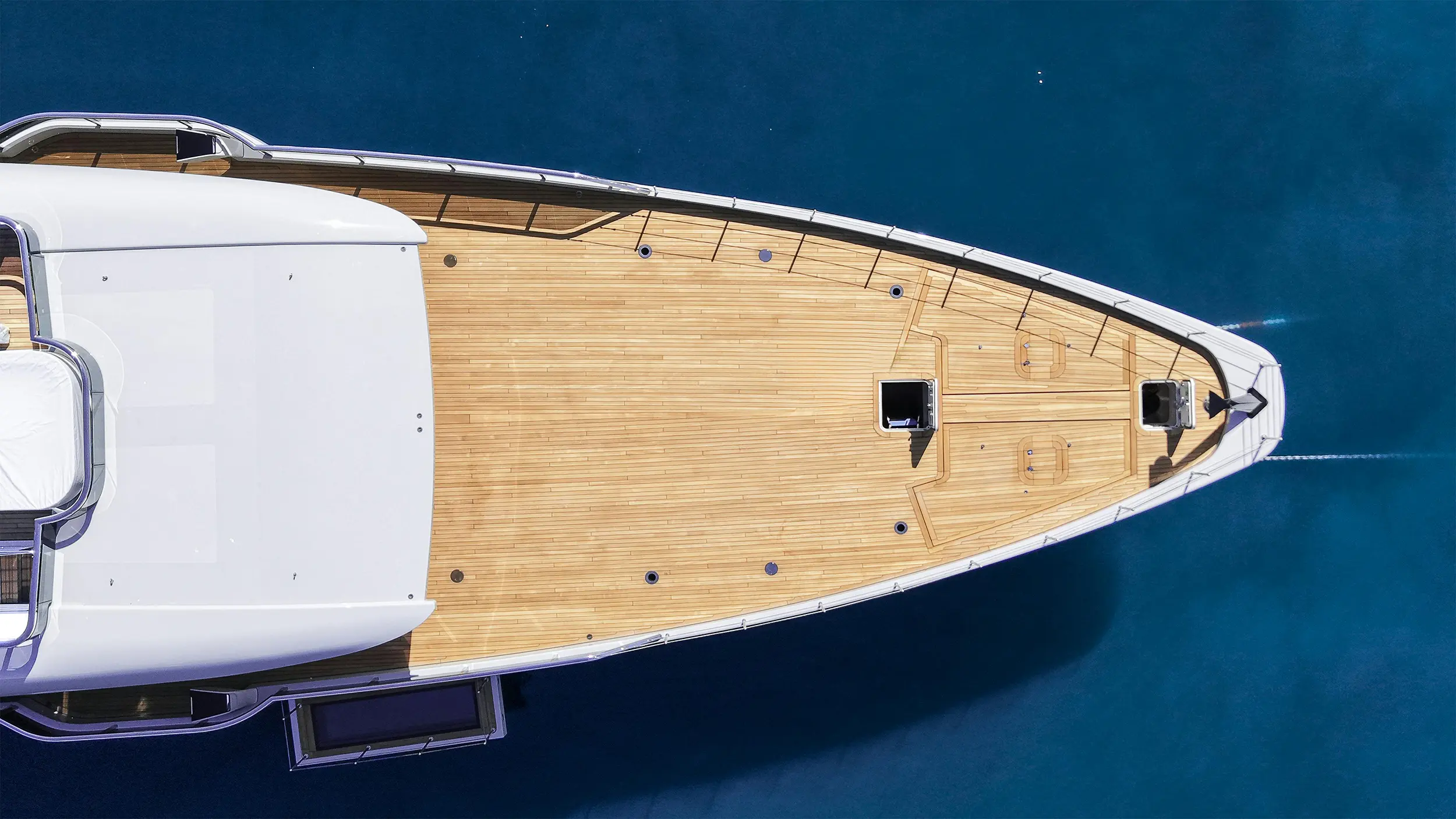 Aerial view of the wooden deck and bow of a white yacht floating on blue water.